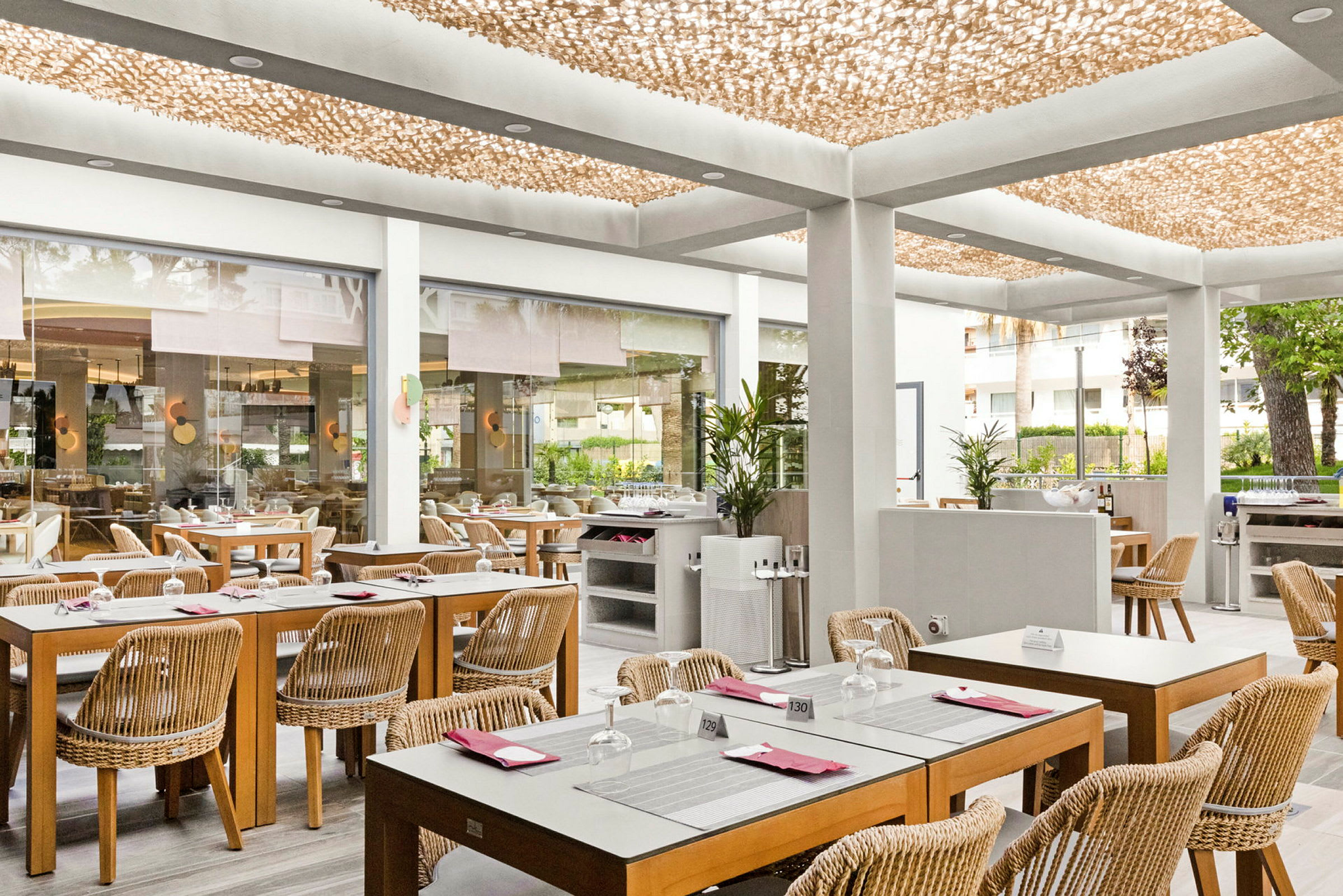 En lys restaurant med trebord og rottingstoler, plassert i et moderne innemiljø med et dekorativt tak og glassvegger som åpner opp til utsiden. (AI-generert tekst)