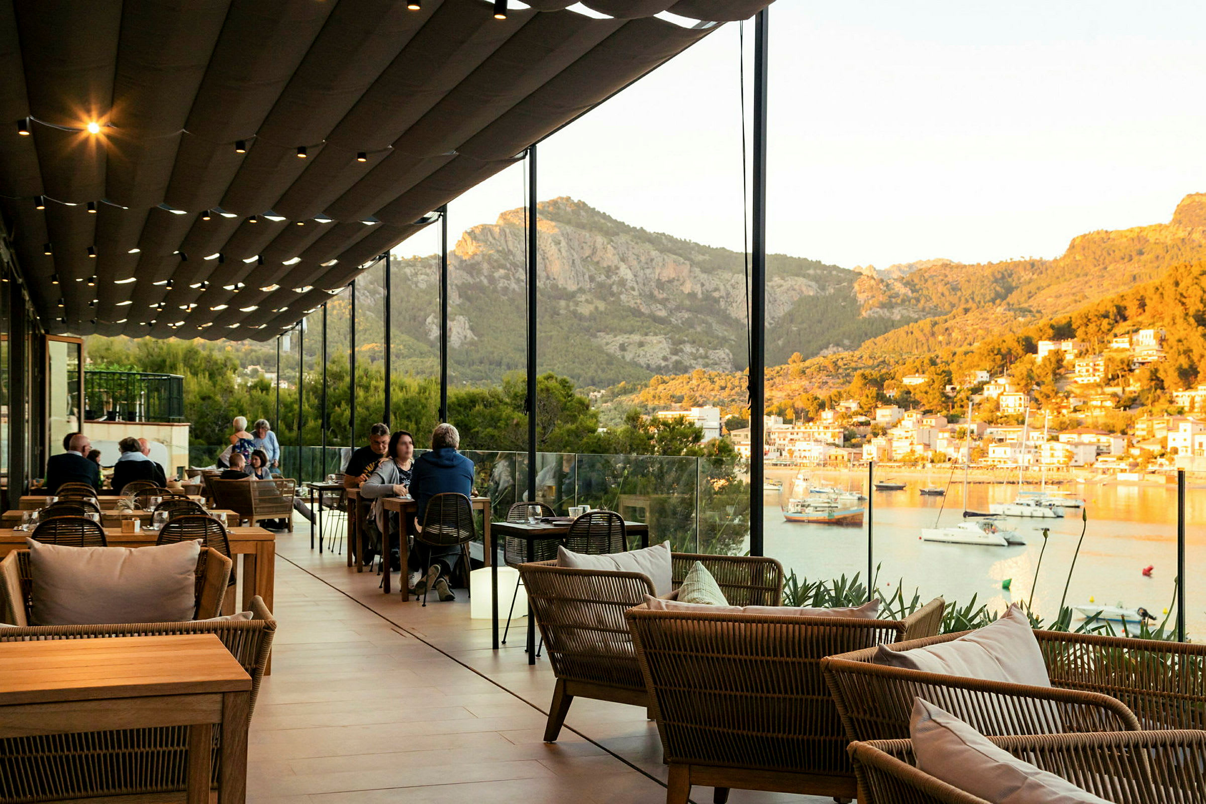 En restaurant med utendørs sitteplasser under et dekke med utsikt over en havn og fjell i bakgrunnen. (AI-generert tekst)