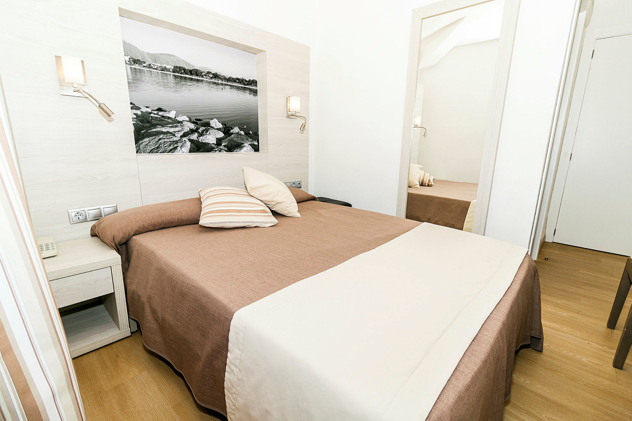 Et hotellrom med en seng dekket med brunt og hvitt sengetøy, med et nattbord ved siden av og et speil som reflekterer rommet. (AI-generert tekst)