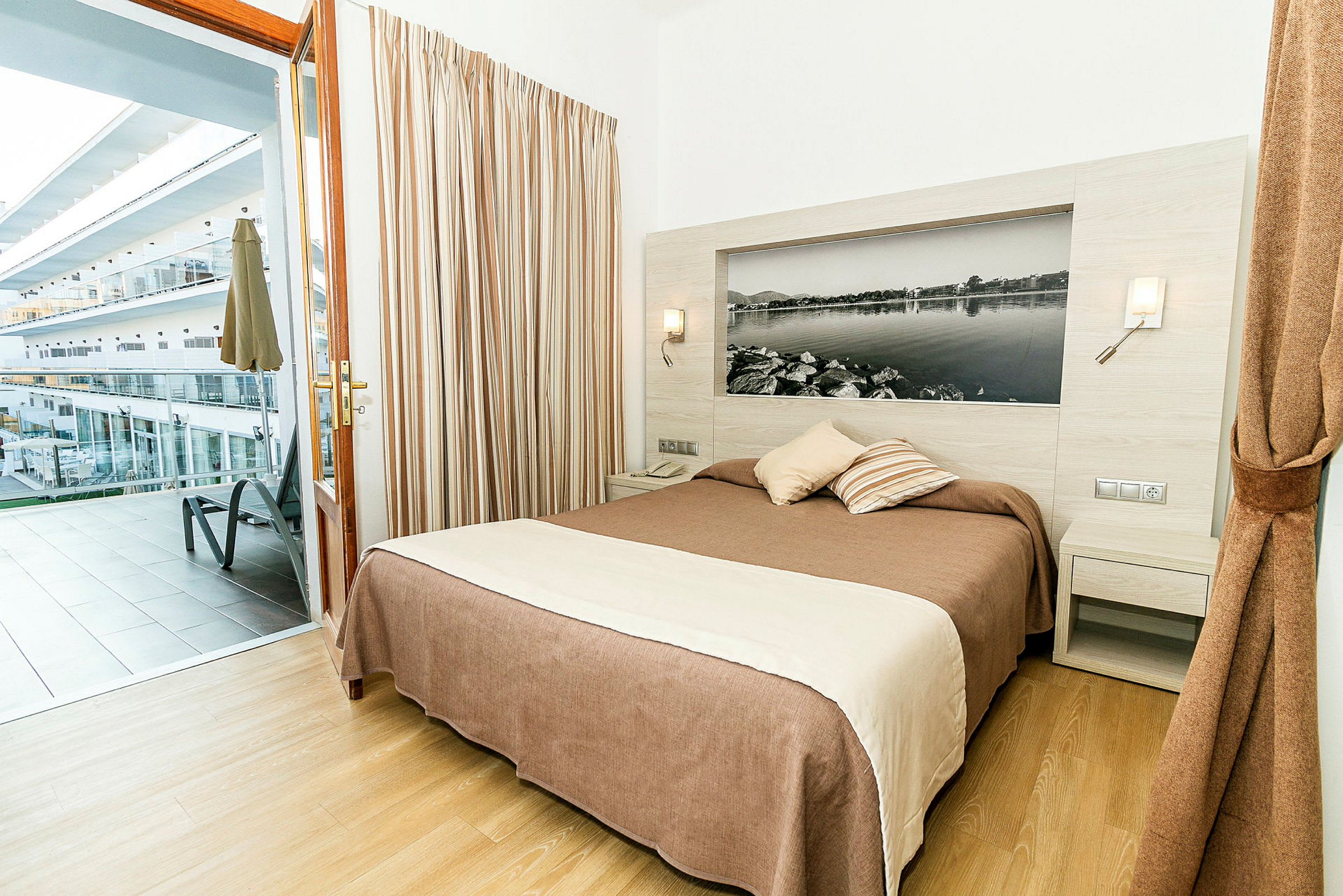 Et hotellrom med en dobbeltseng og utgang til en balkong med utsikt over andre bygninger. (AI-generert tekst)