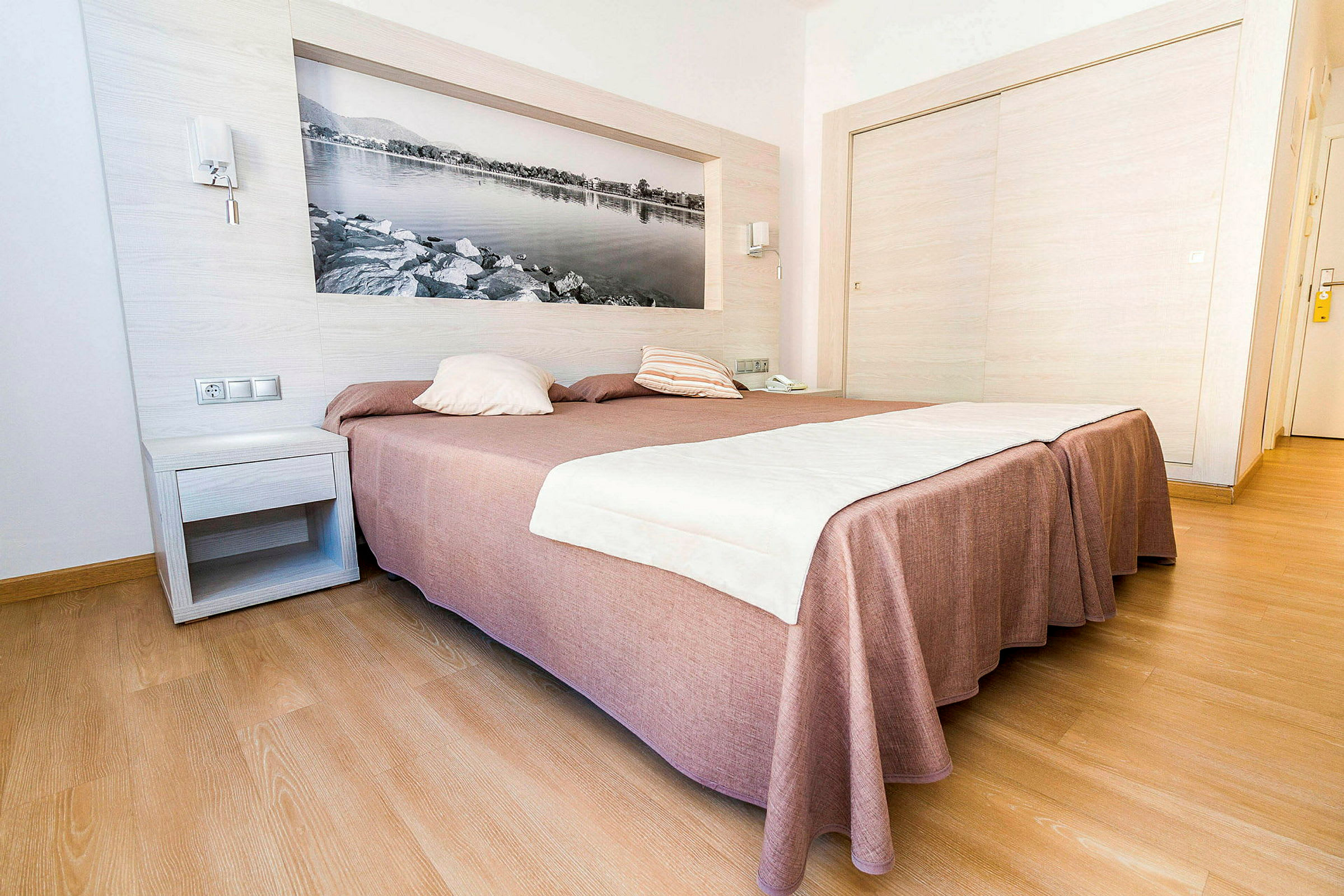 Hotellrom med en stor seng dekket med et brunaktig sengeteppe og hvitt sengetøy, med tregulv og en veggdekorasjon i form av et svart-hvitt bilde. (AI-generert tekst)