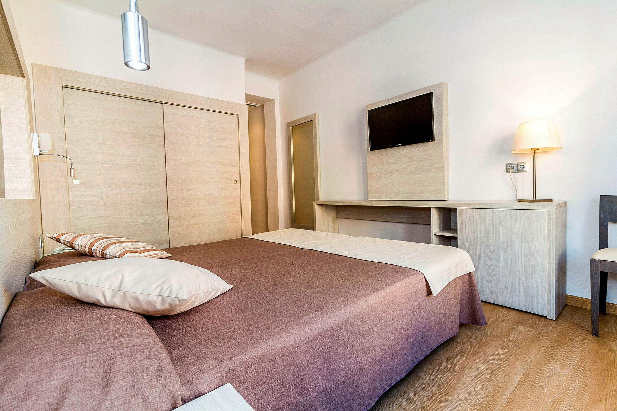 Et hotellrom med en dobbeltseng dekket med brun sengeteppet og hvite puter. På veggen henger en flatskjerm-TV over et tre-skrivebord. Rommet er lyst og moderne innredet. (AI-generert tekst)