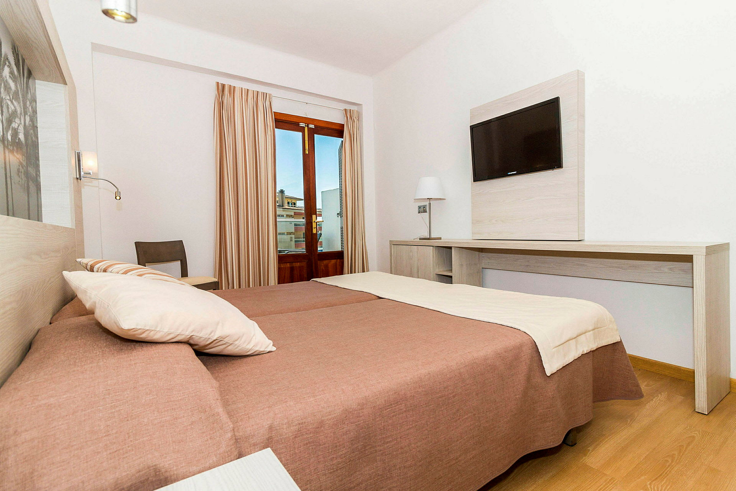 Et hotellrom med to senger, skrivebord og veggmontert TV samt et vindu med beige gardiner. (AI-generert tekst)