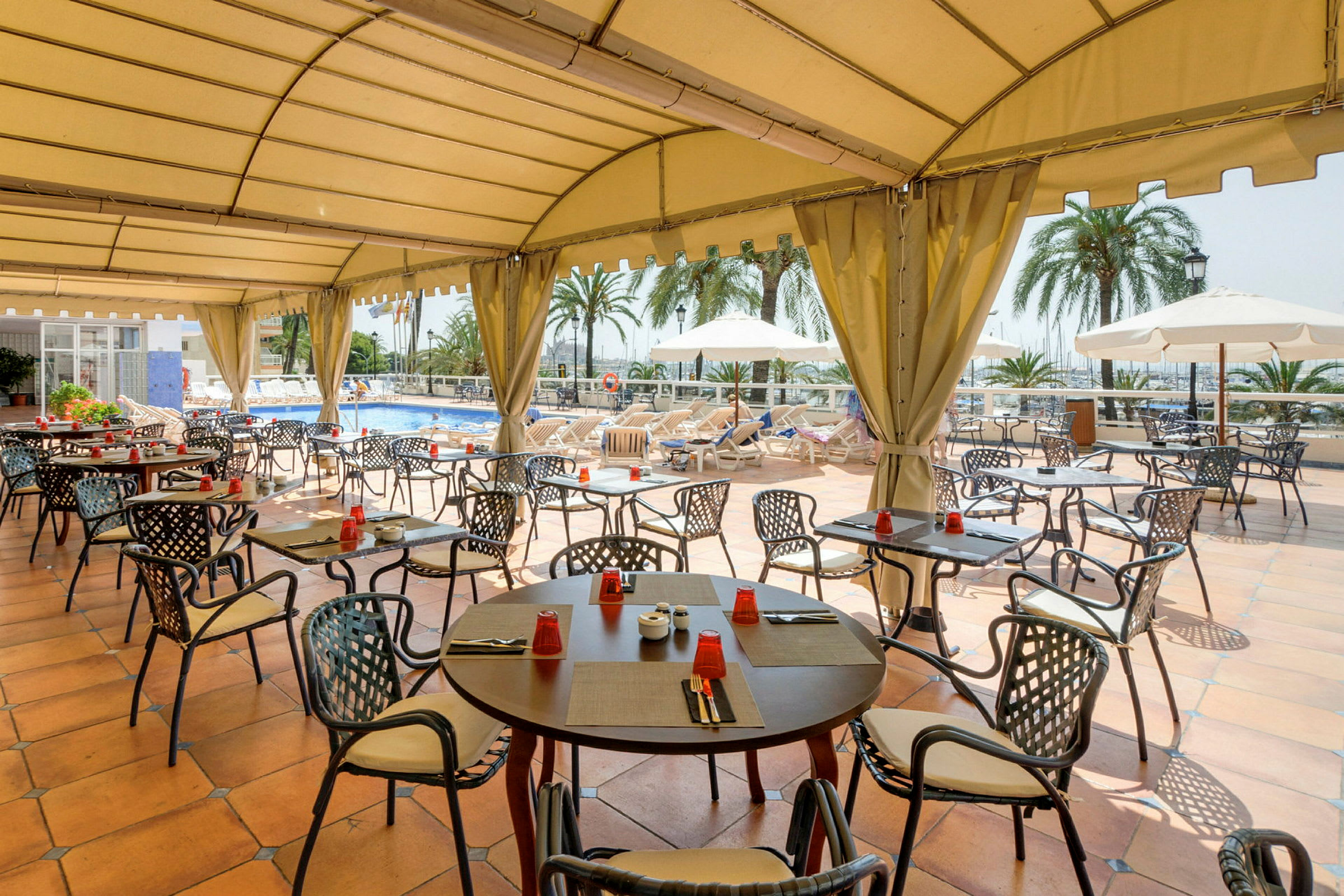 En restaurant terrasse med tak, der bord og stoler er plassert rundt et basseng. (AI-generert tekst)
