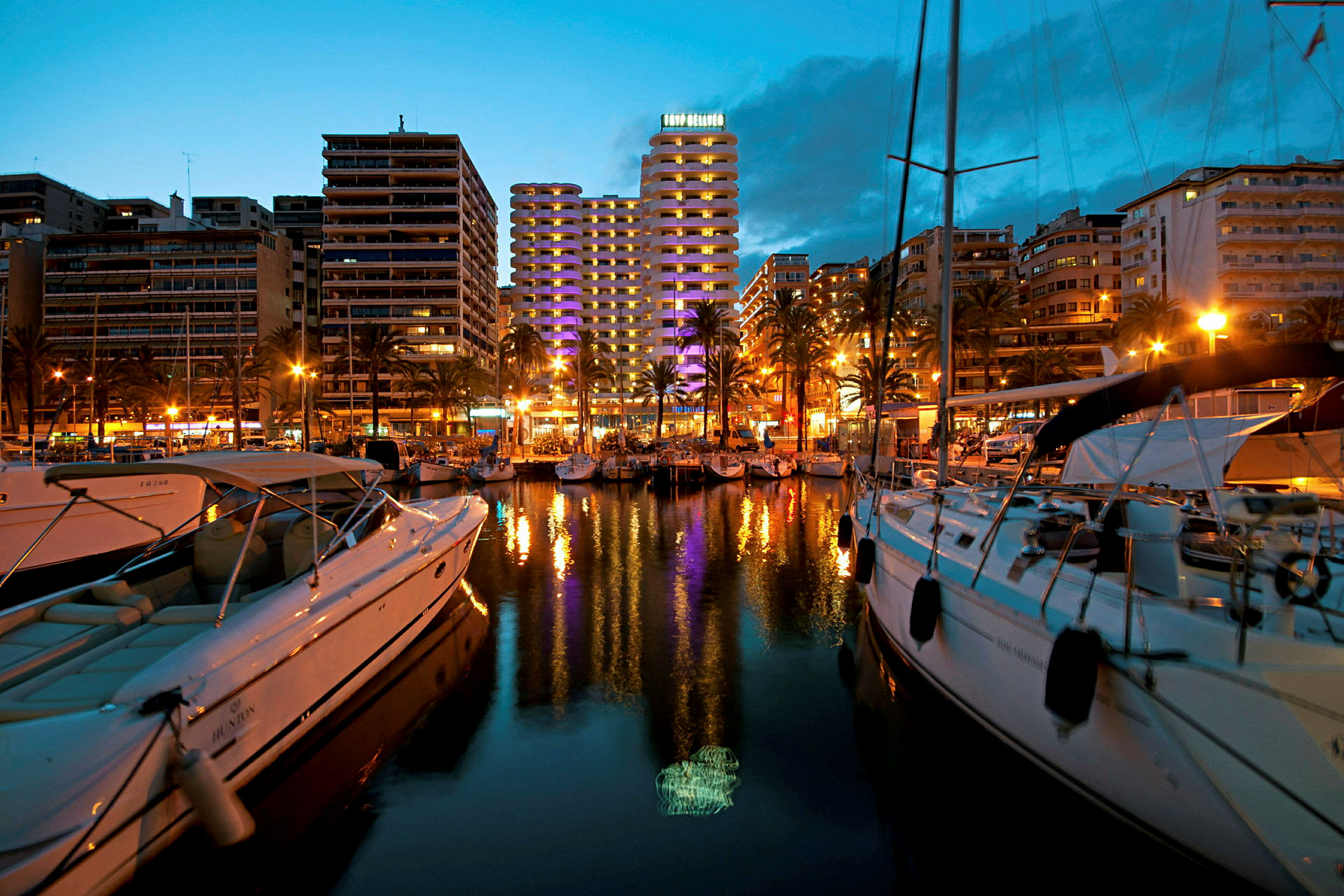 En marina i en by omgitt av store bygninger med belysning, båter og vann som reflekterer lysene. (AI-generert tekst)