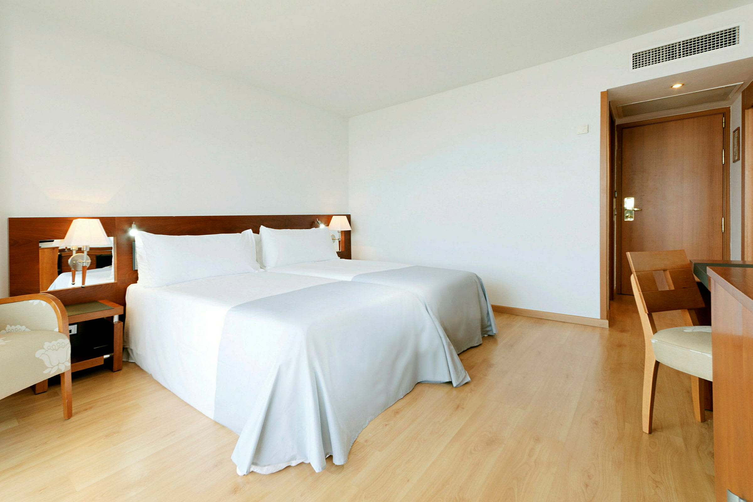 Et hotellrom med to senger, hvitt sengetøy og en trepanel bak sengene. Det er et skrivebord med en stol og en dør i bakgrunnen. (AI-generert tekst)