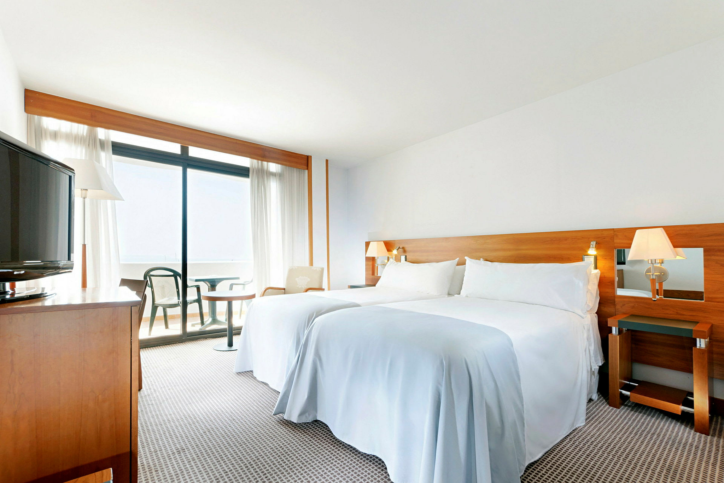 Et lyst, romslig hotellrom med en dobbeltseng, tremøbler, en balkong og utsikt gjennom store vinduer. (AI-generert tekst)