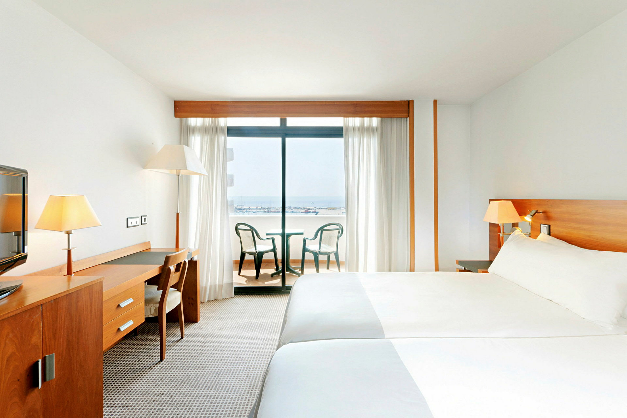 Hotellrom med en stor seng, et skrivebord med stol, og balkongmøbler foran en glassdør som viser en utsikt over havet. (AI-generert tekst)