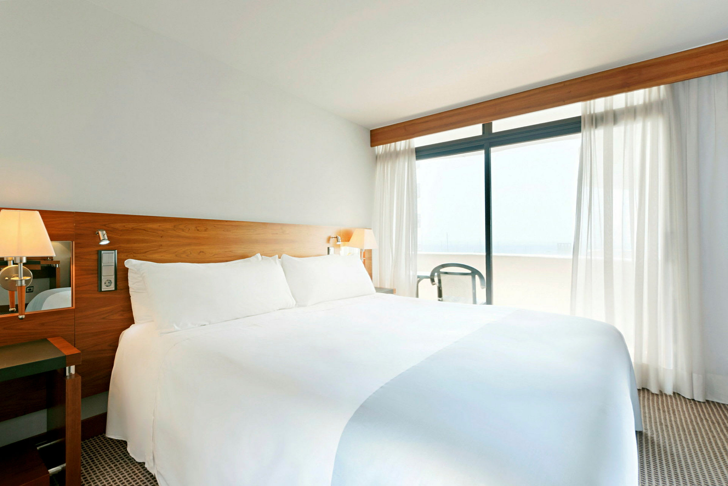 Et lyst hotellrom med en stor oppredd seng med hvitt sengetøy, en hodegavl i tre og vinduer med gardiner som leder ut til en balkong. (AI-generert tekst)