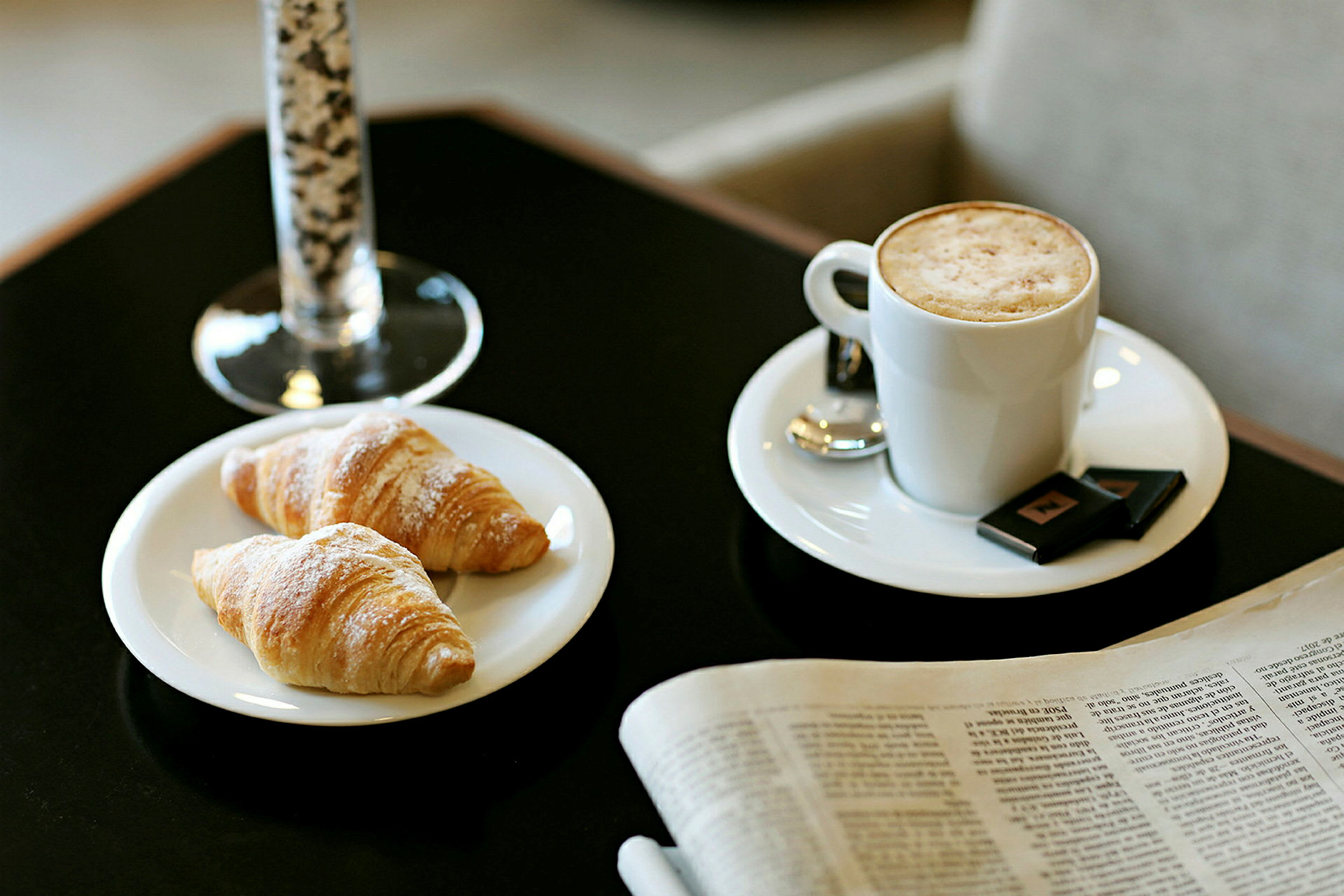 Bildet viser to croissanter på en tallerken, en kopp kaffe med melkeskum og et glass i bakgrunnen. En avis ligger ved siden av på bordet. (AI-generert tekst)