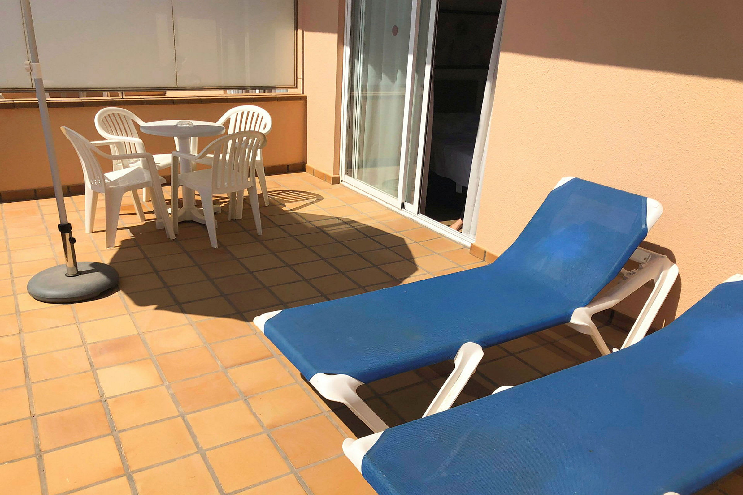 Terrasse med to solsenger, et hvitt bord og fire hvite stoler, samt en parasoll. (AI-generert tekst)
