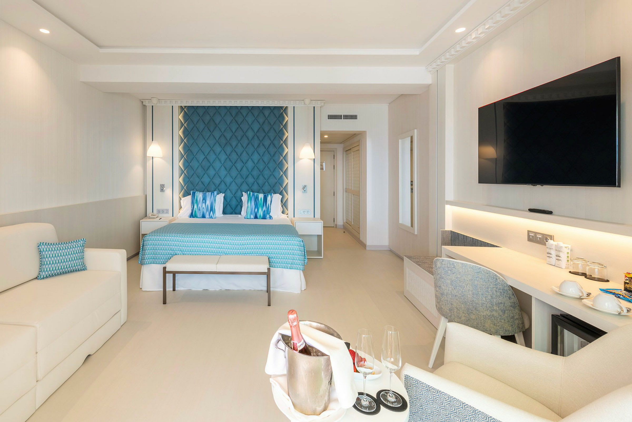 Et hotellrom med en dobbeltseng, blå sengegavl og hvite møbler, samt et bord med champagne og glass. (AI-generert tekst)
