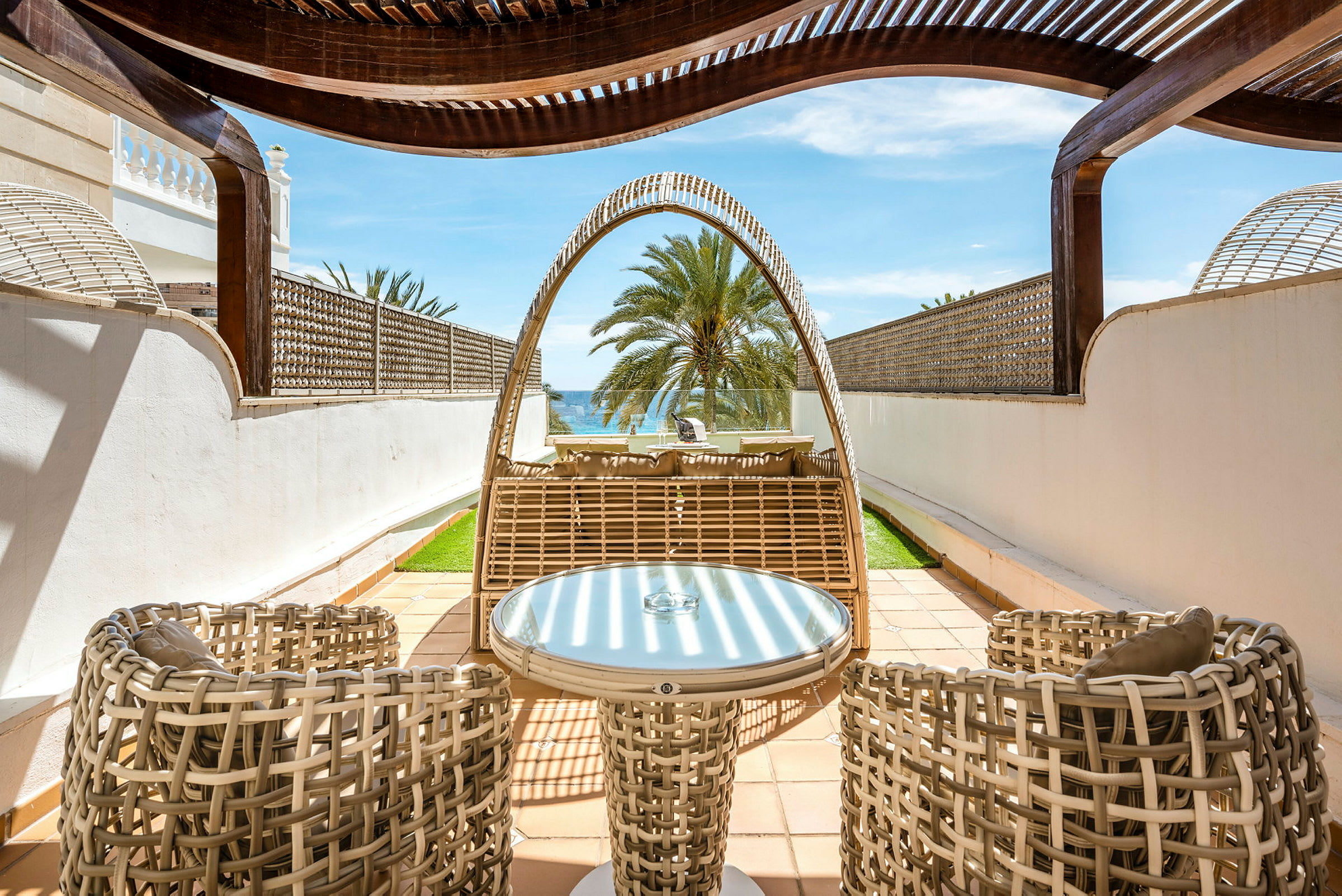 Utemøbler i flettet design på en privat terrasse med utsikt over en palme og havet. (AI-generert tekst)