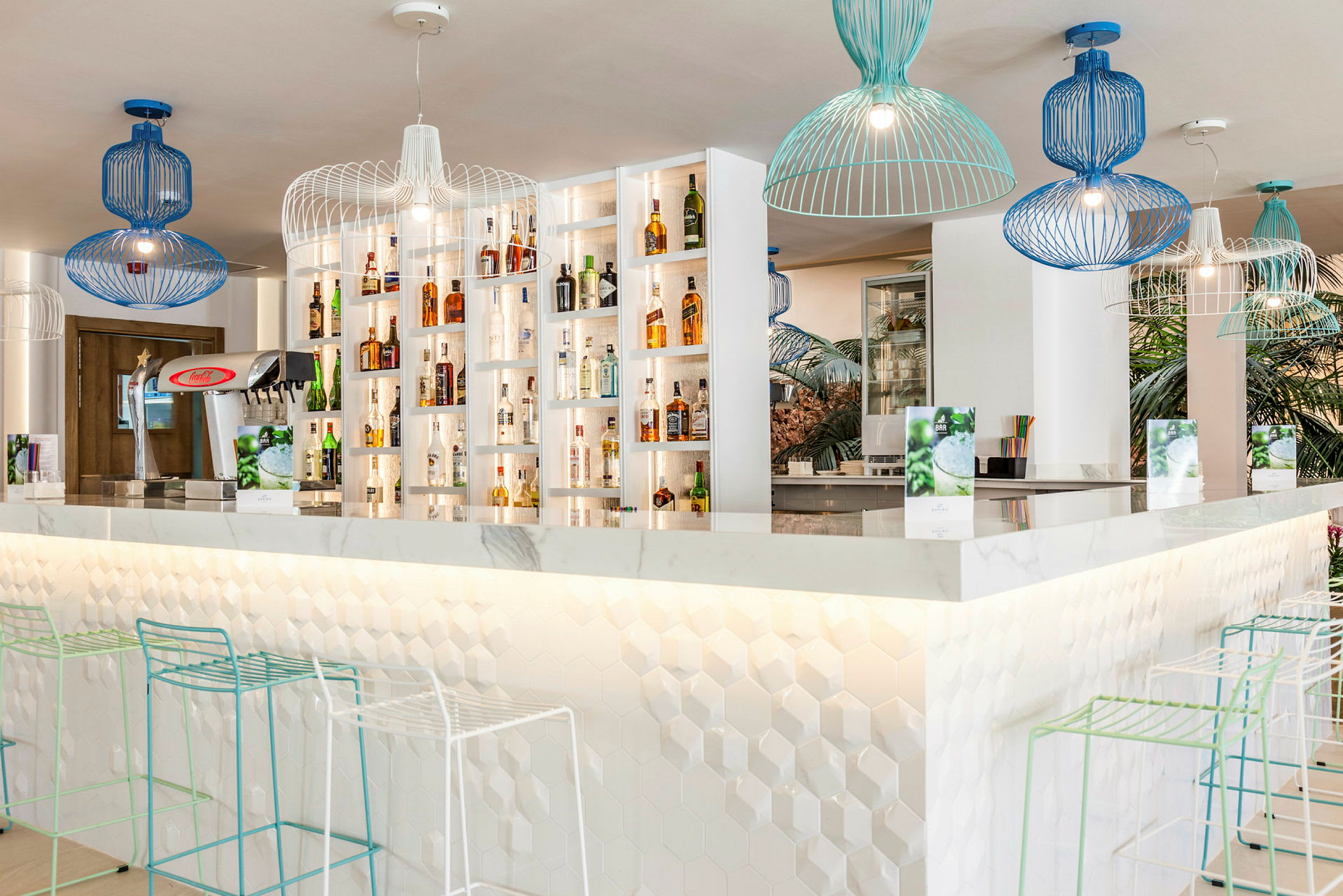 En moderne bar med hvite hyller fylt med flasker, geometriske lamper i blått og grønt henger fra taket, og stoler med smale metallrammer står ved bardisken. (AI-generert tekst)
