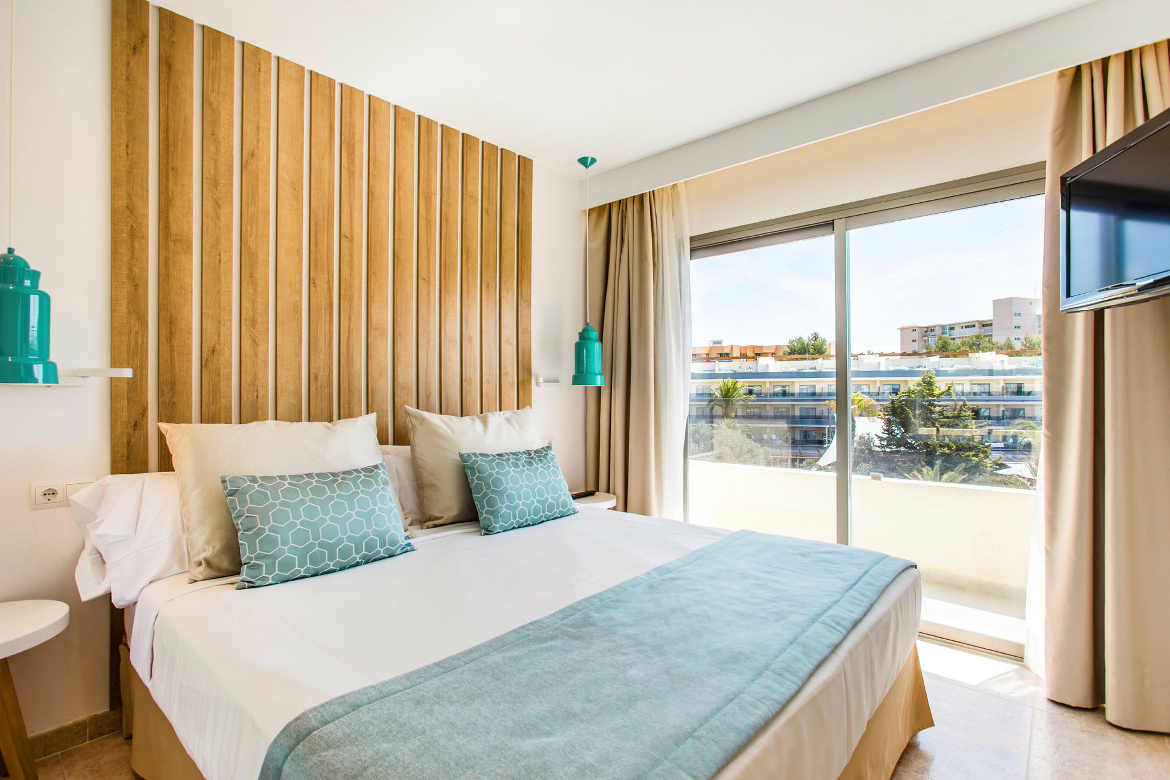 Et moderne hotellrom med en stor seng, trepanel bak sengen, hvite og blå sengetøy, samt en vindusutsikt over bygninger og trær utenfor. (AI-generert tekst)
