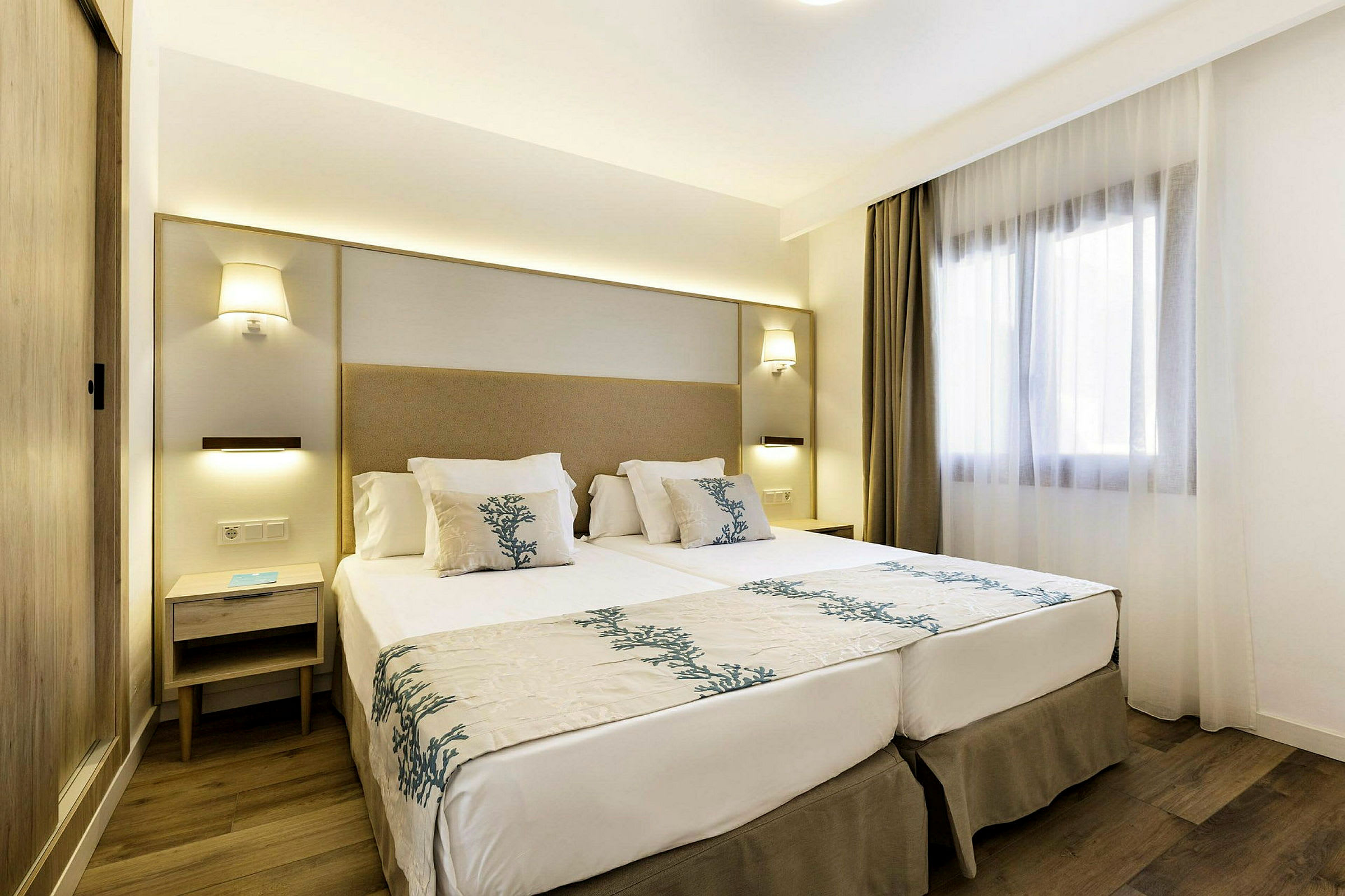 Moderne innredet hotellrom med en dobbeltseng, nattbord og et vindu med lyse gardiner. (AI-generert tekst)