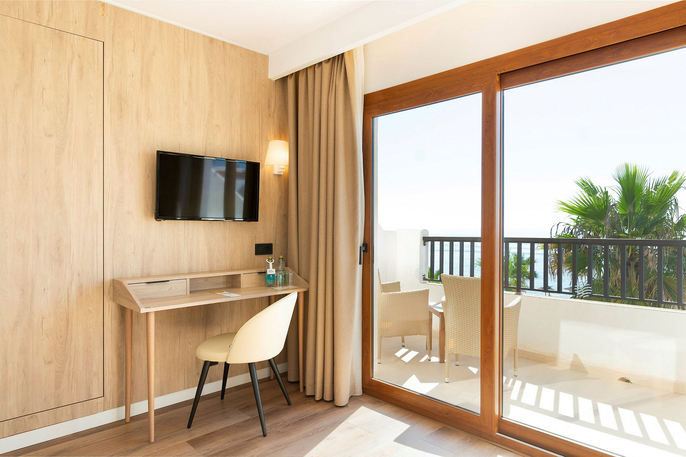 Hotellrom med trepanel, skrivebord, stol og en TV på veggen. Store vinduer med utsikt mot en balkong med møbler og palmer. (AI-generert tekst)