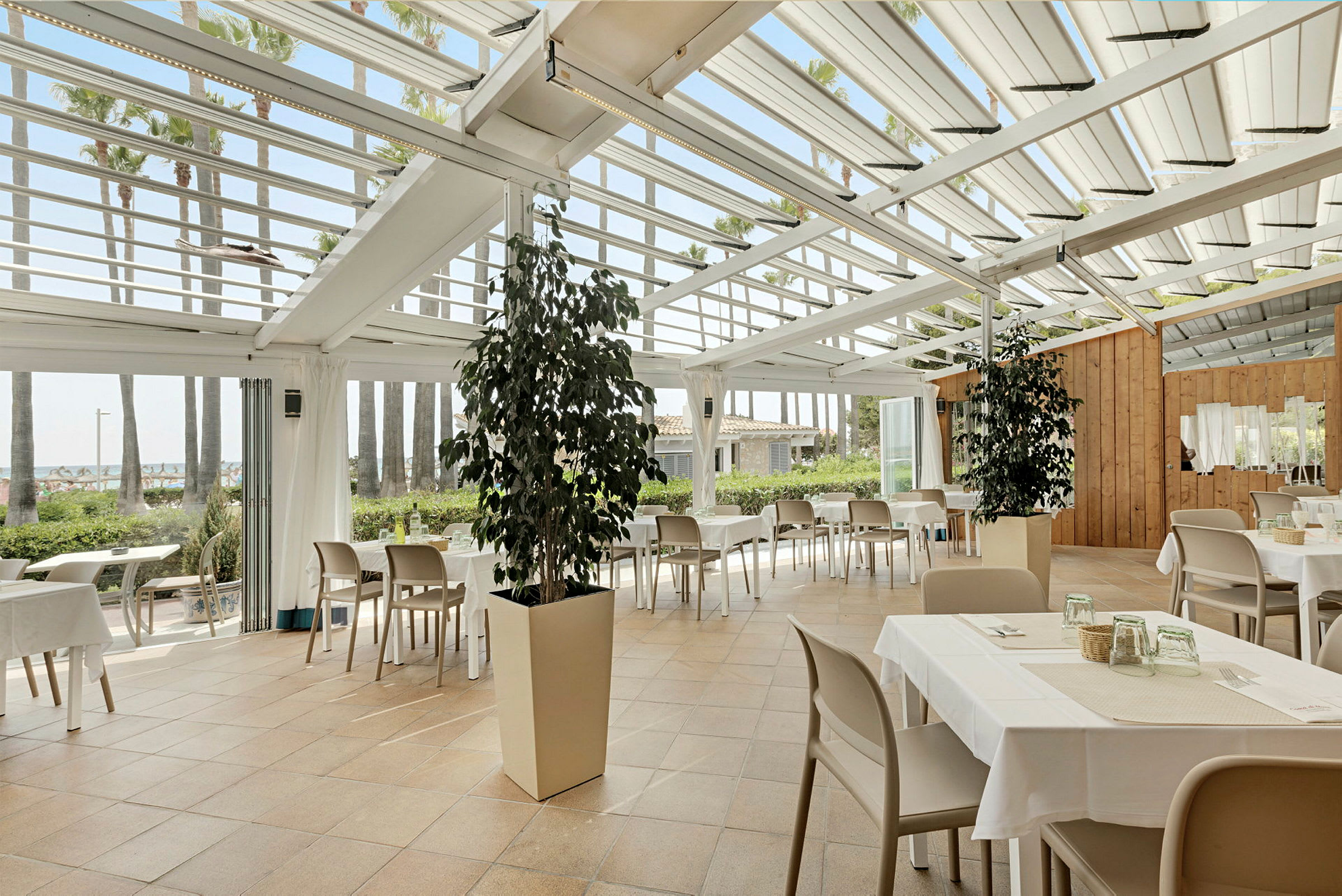 Restaurant med bord og stoler under et glasstak, omgitt av grønne planter og palmer. (AI-generert tekst)