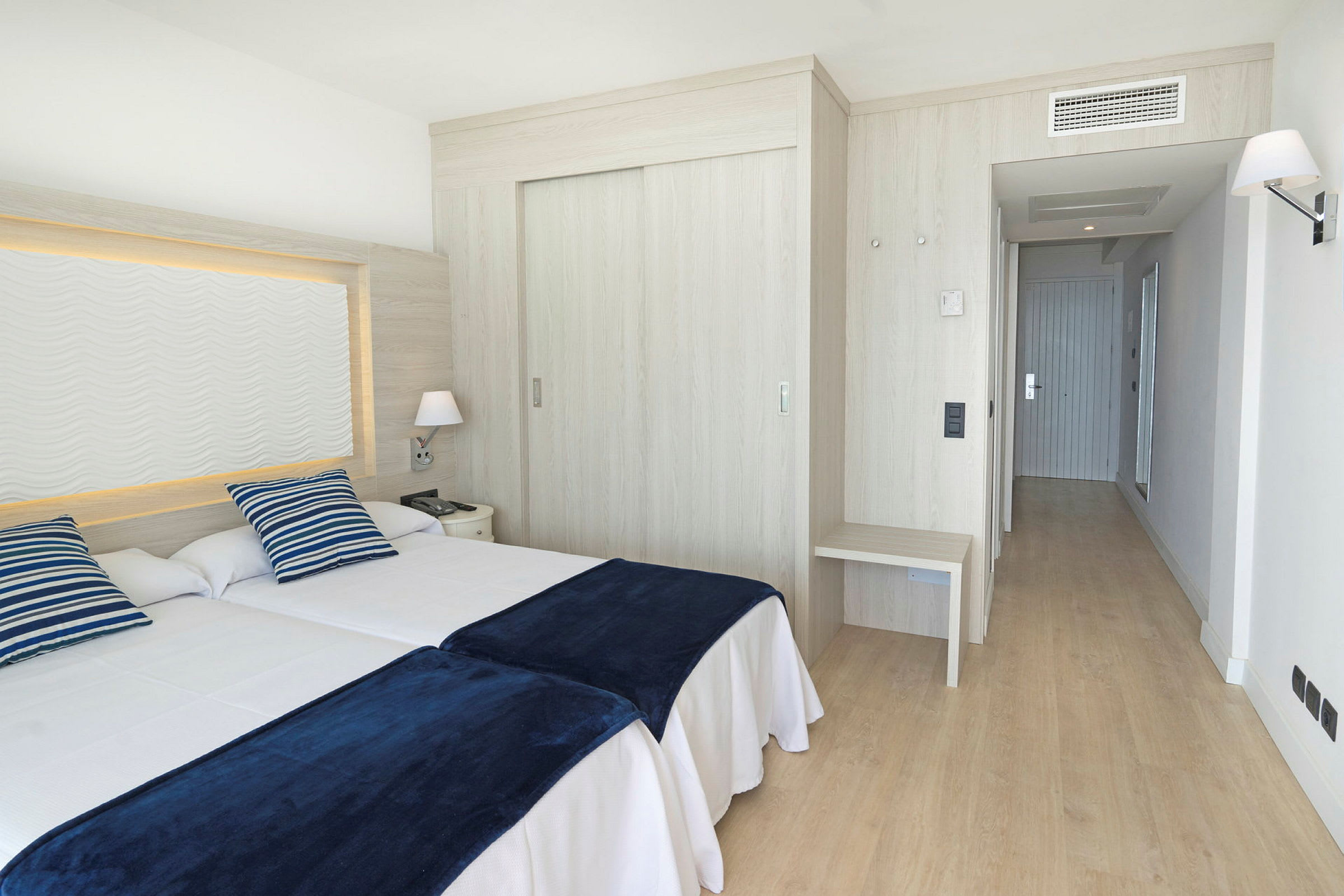 Et hotellrom med to enkeltsenger, blåstripete puter og mørkeblå tepper. (AI-generert tekst)