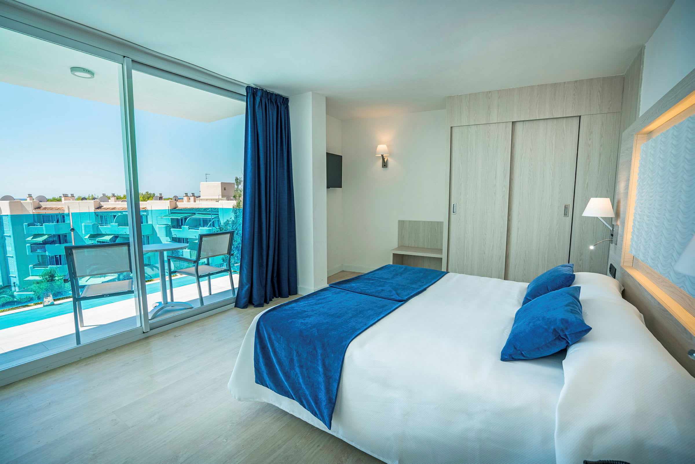 Et hotellrom med en stor seng, blått sengeteppe og puter. Rommet har også et stort glassvindu som vender mot en balkong med utsikt over blå bygninger. (AI-generert tekst)