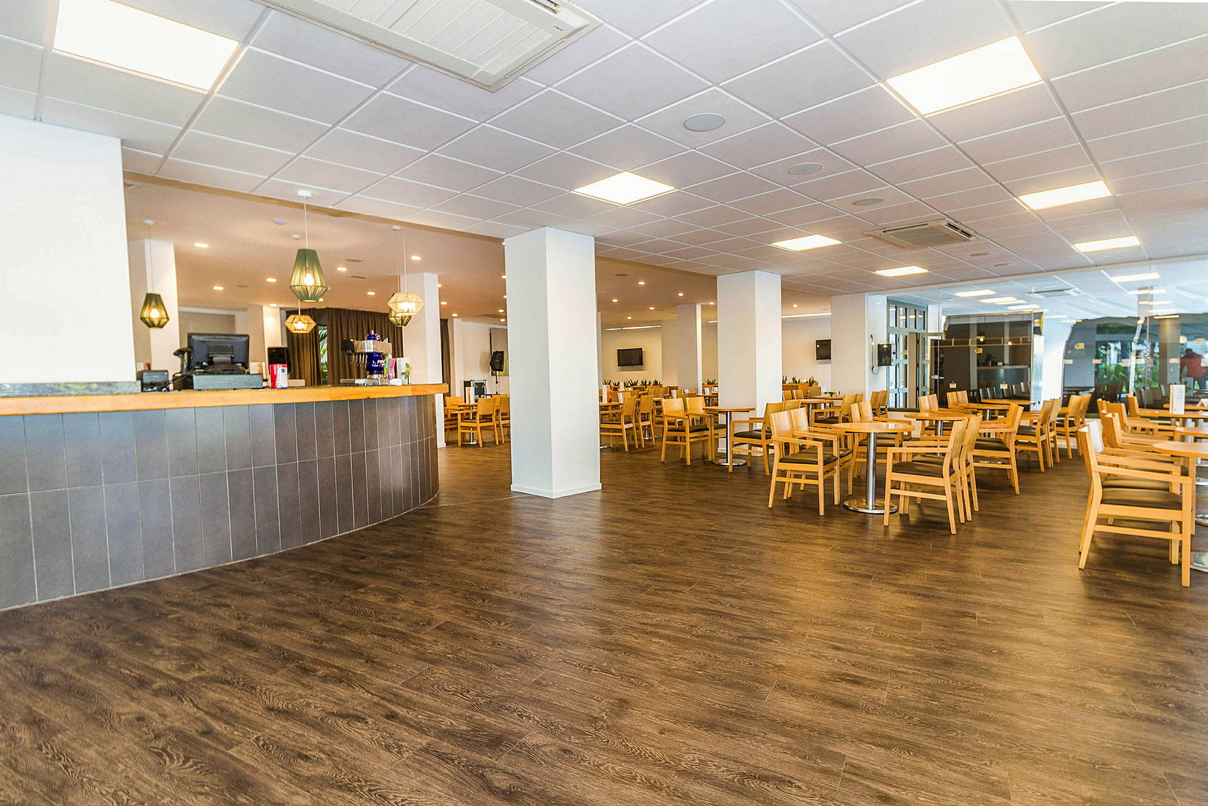 Et romslig kafé- eller restaurantområde med lyse bord og stoler i tre plassert på et mørkt gulv. I forgrunnen er det en bar med hengende lamper over. (AI-generert tekst)