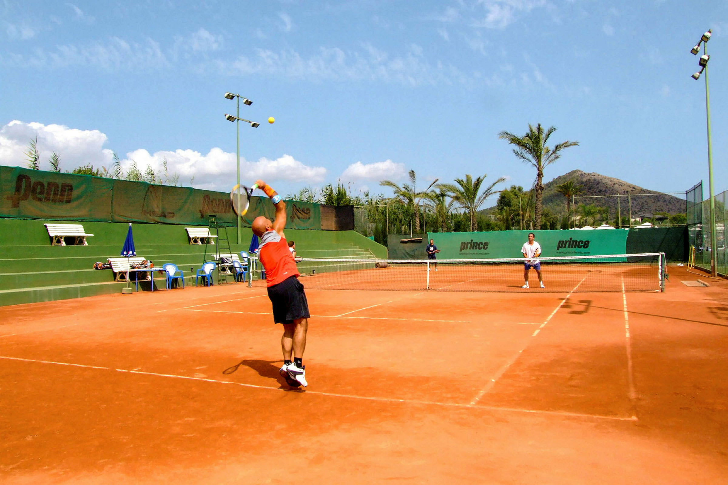 To personer spiller tennis på en grusbane utendørs på en solrik dag. (AI-generert tekst)