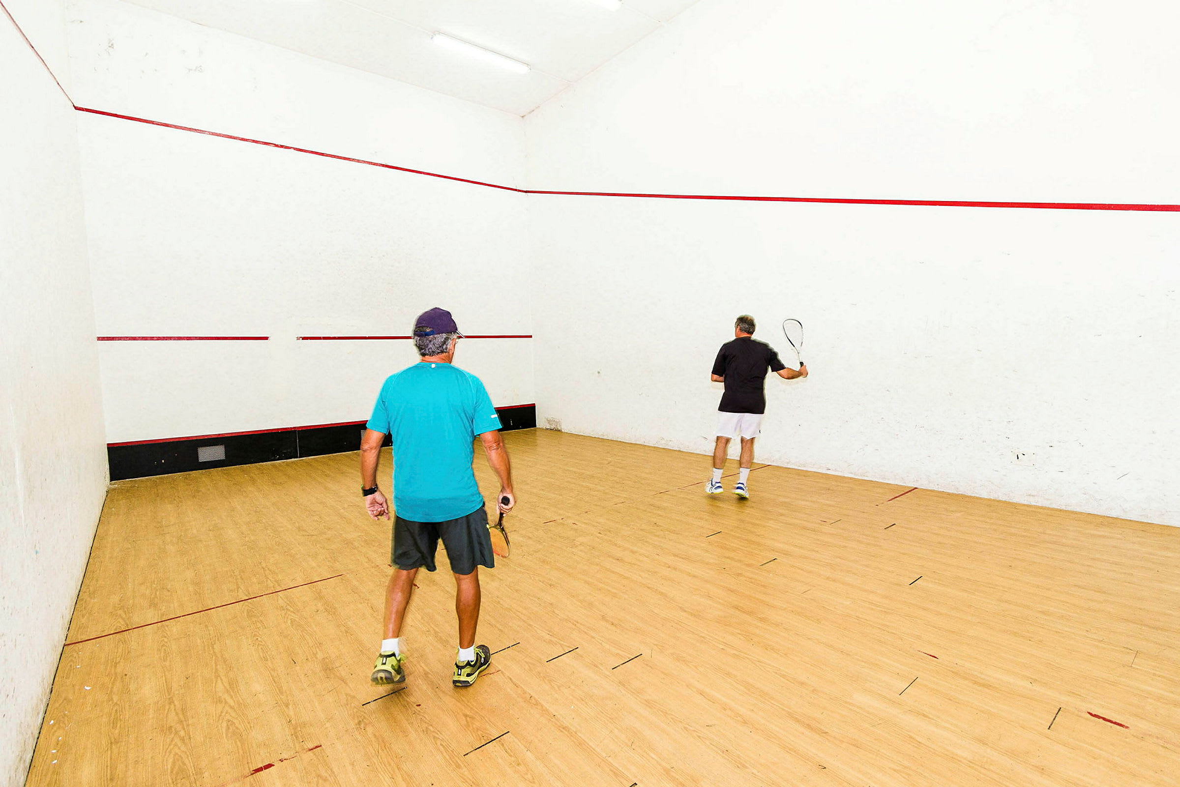 To personer spiller squash på en innendørsbane med lyse vegger og tregulv. (AI-generert tekst)