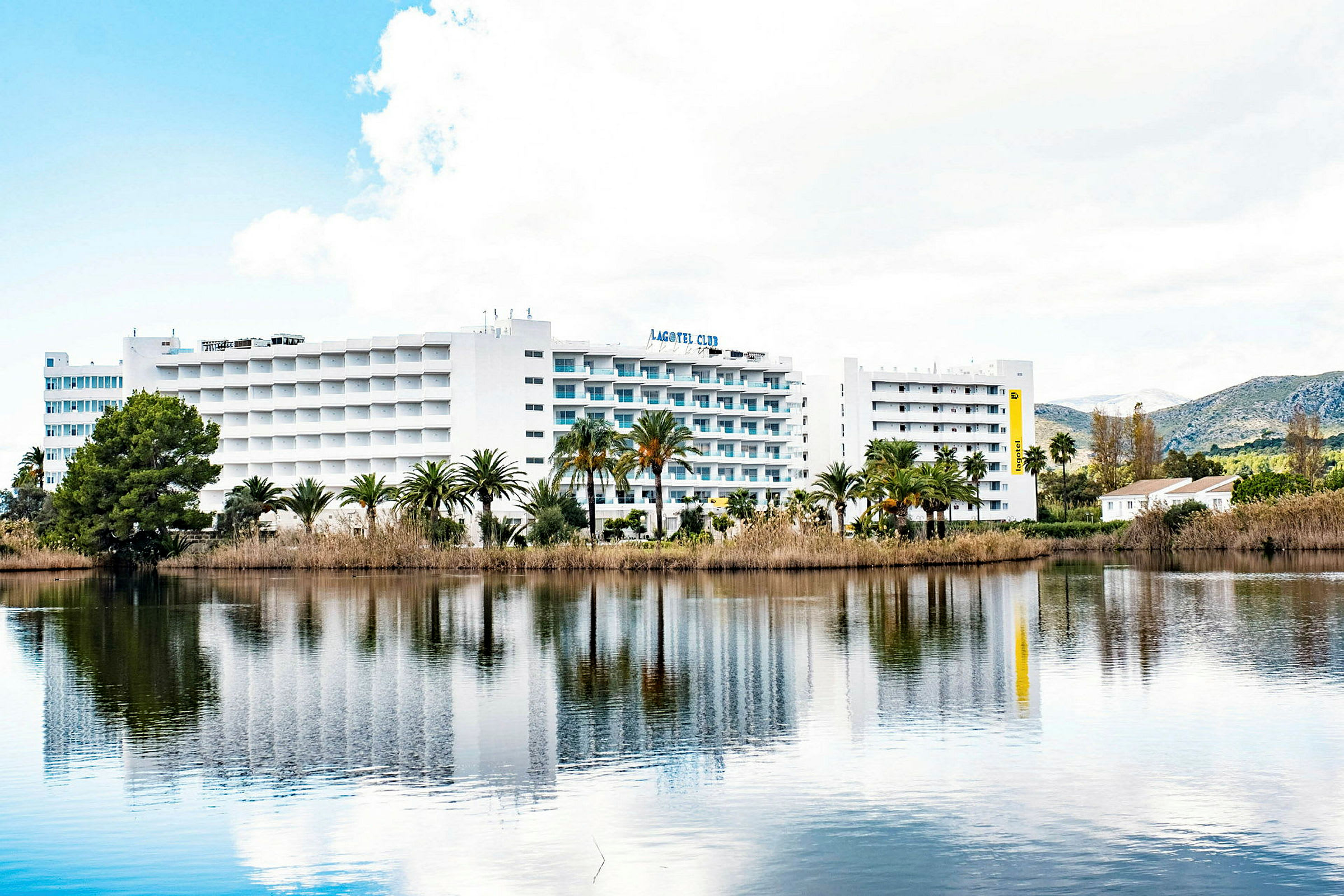Et hotellkompleks med hvit fasade og balkonger reflekteres i en stille innsjø. Grønne palmer og vegetasjon omgir bygningen. (AI-generert tekst)