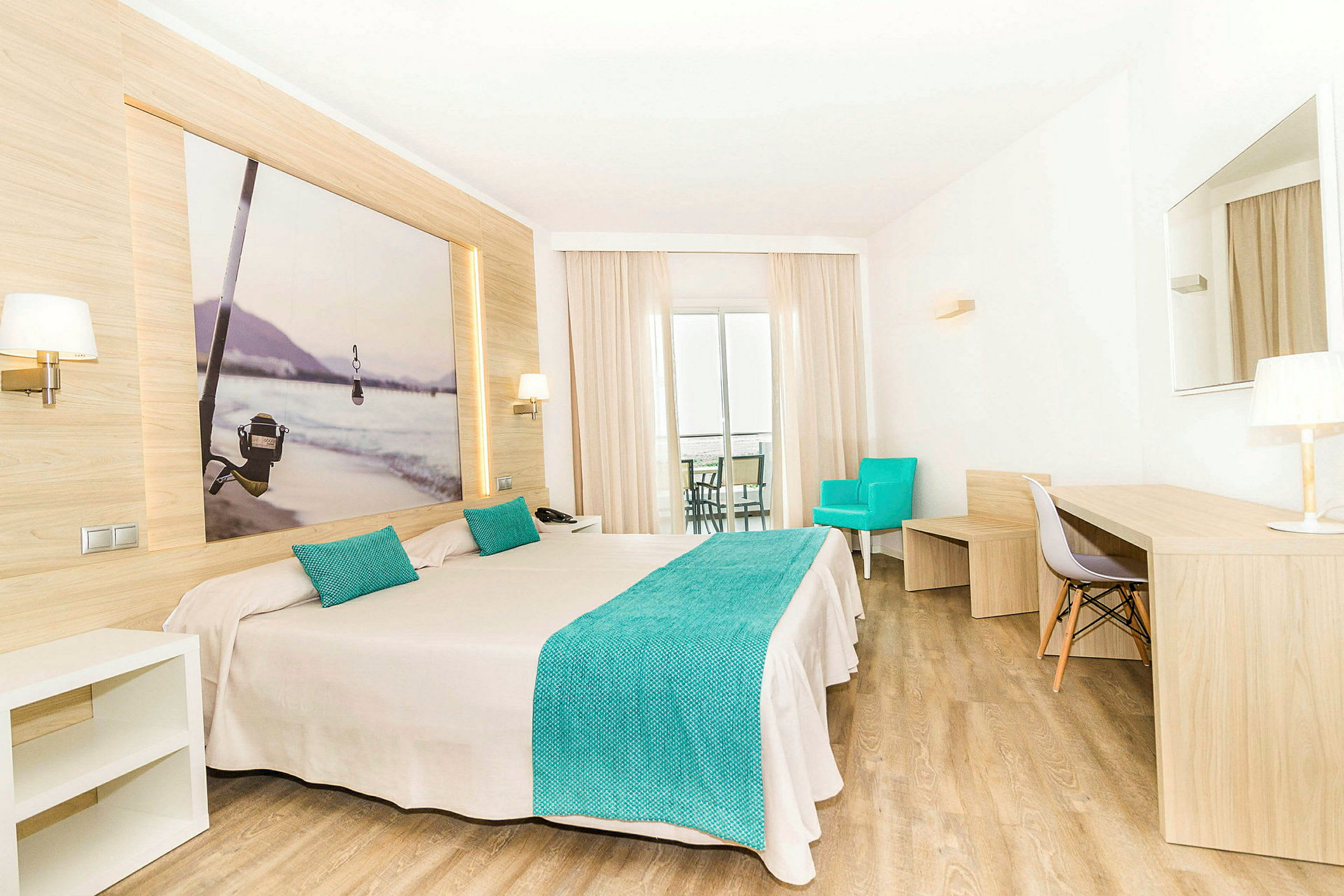 Et hotellrom med en dobbeltseng, turkise detaljer og moderne tredesign. Bak sengen henger et bilde med et fiske-tema. Rommet har et skrivebord, en turkis lenestol og en balkong. (AI-generert tekst)