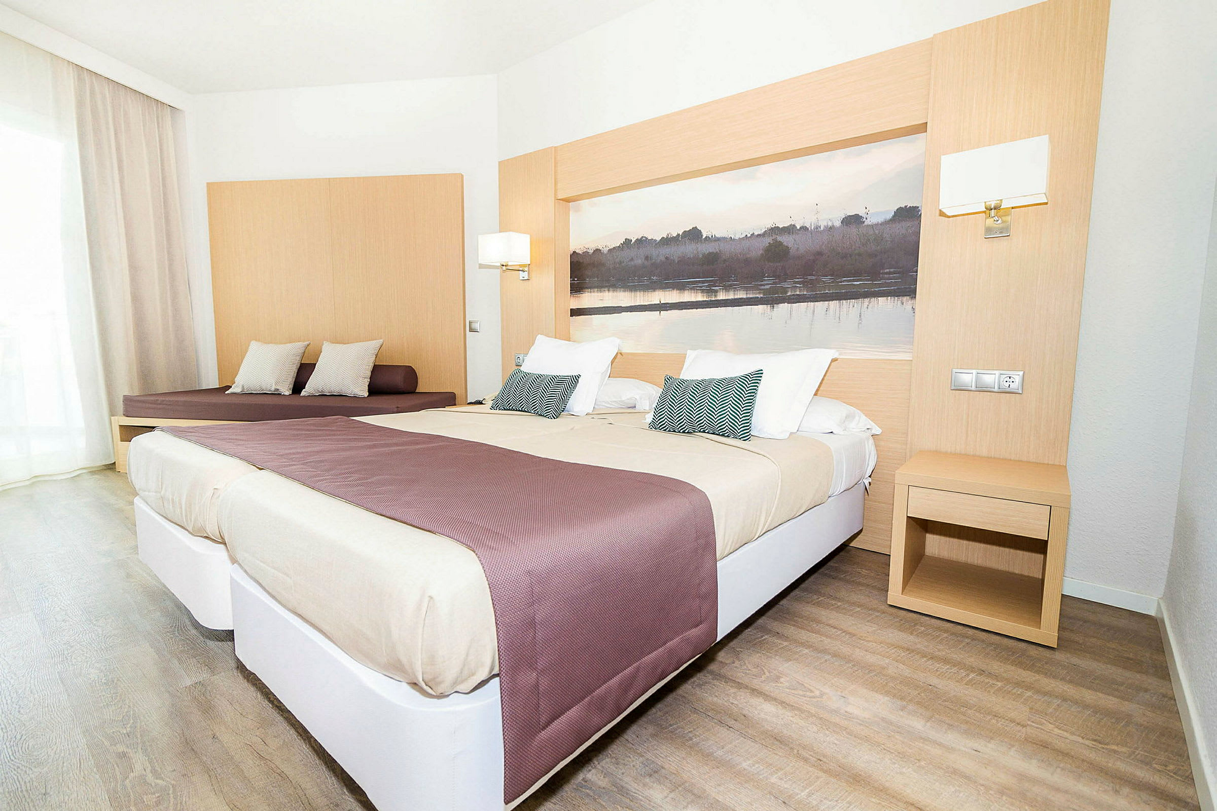 Et lyst hotellrom med to senger og en sitteplass. Sengene er oppredd med beige og lilla sengetepper, og en stor bilde dekorerer veggen bak sengene. (AI-generert tekst)