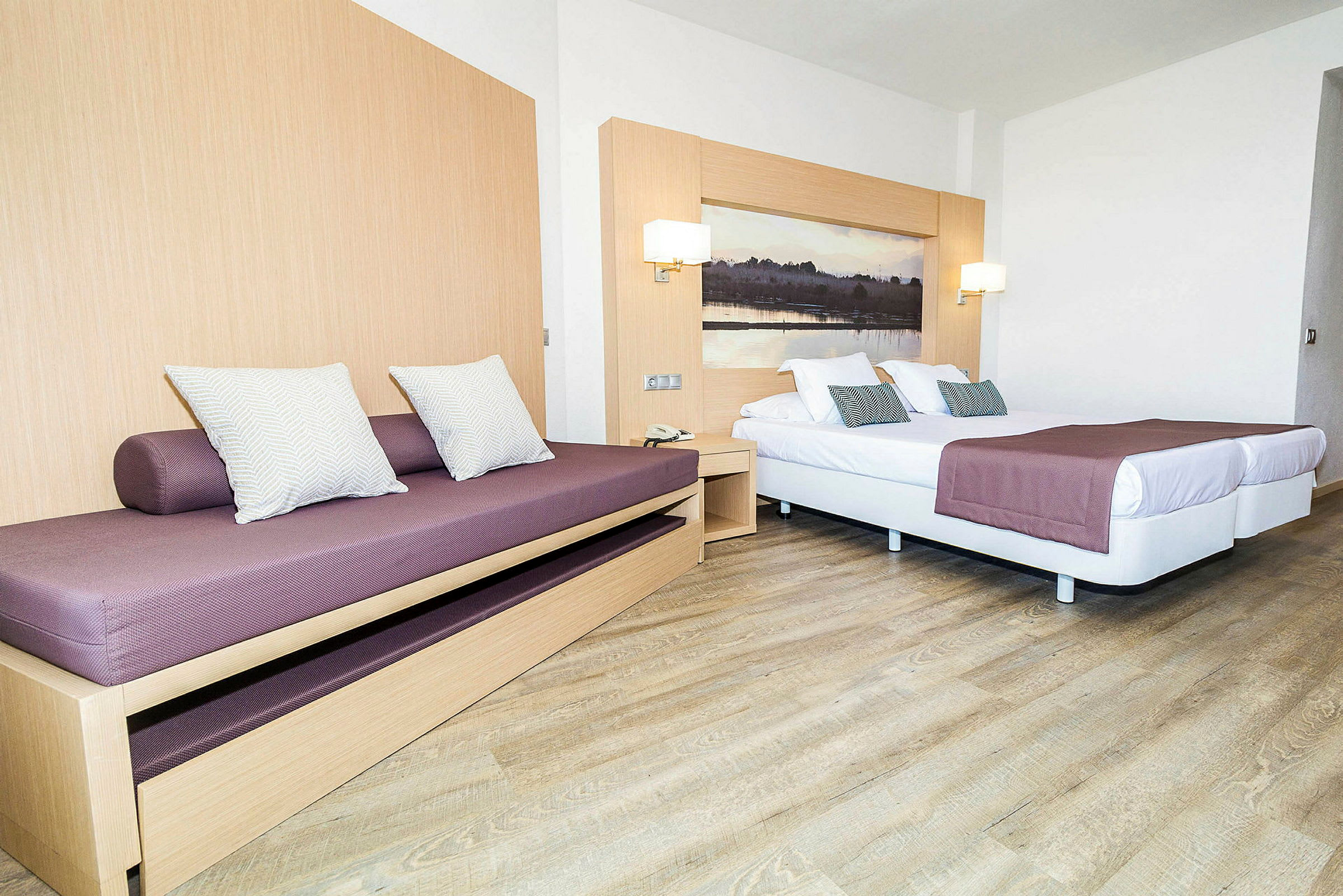 Et hotellrominteriør med en sovesofa og en dobbeltseng med lilla sengetepper og pynteputer. (AI-generert tekst)