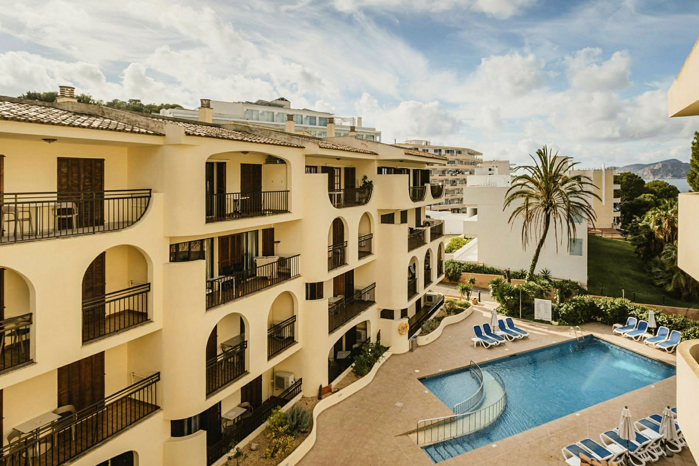 Leilighetsbygg med balkonger rundt et utendørs basseng og en palme. (AI-generert tekst)