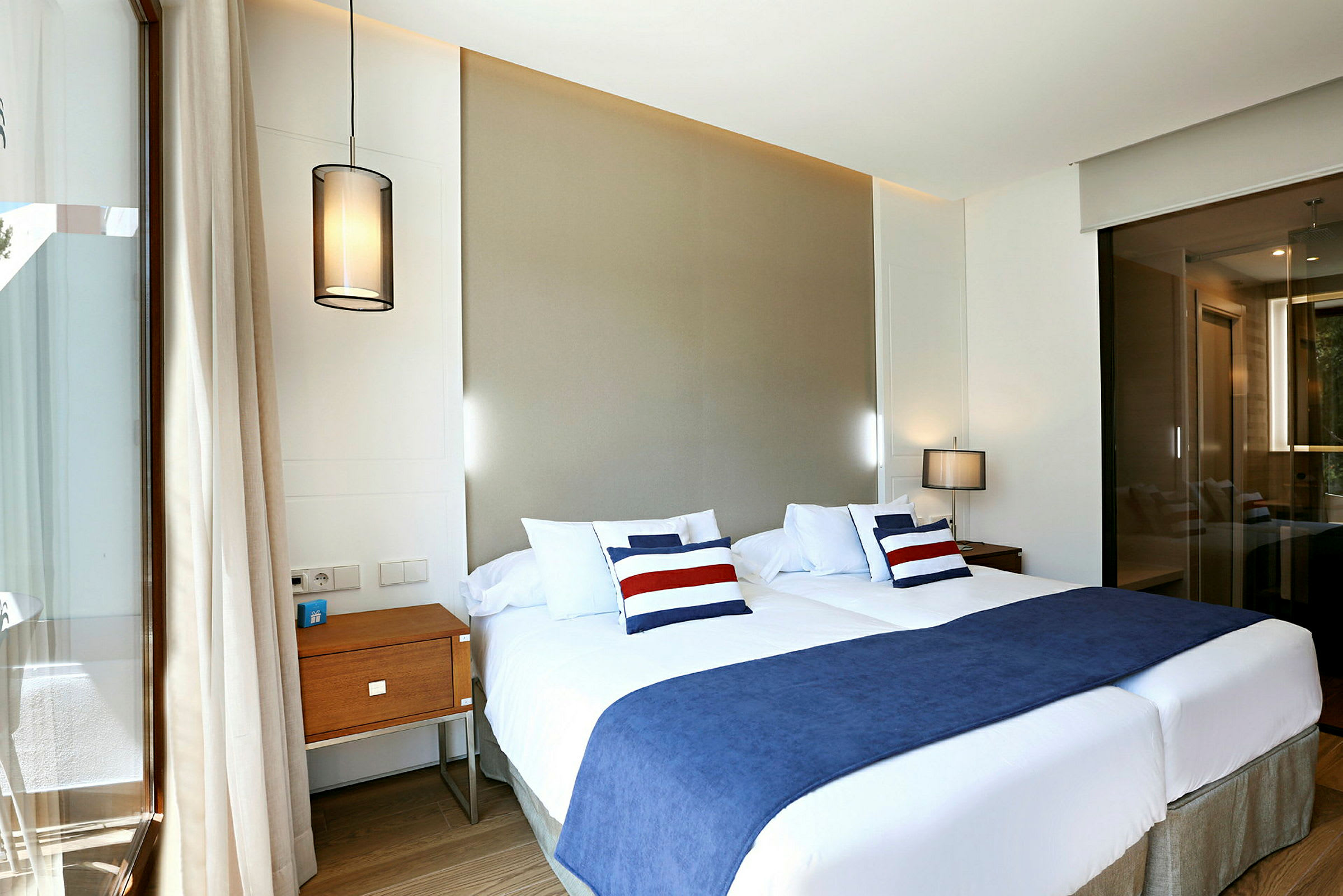 Et hotellrom med to enkeltsenger, blå sengetepper og dekorative puter. Innredningen inkluderer hengende lamper og et nattbord. (AI-generert tekst)