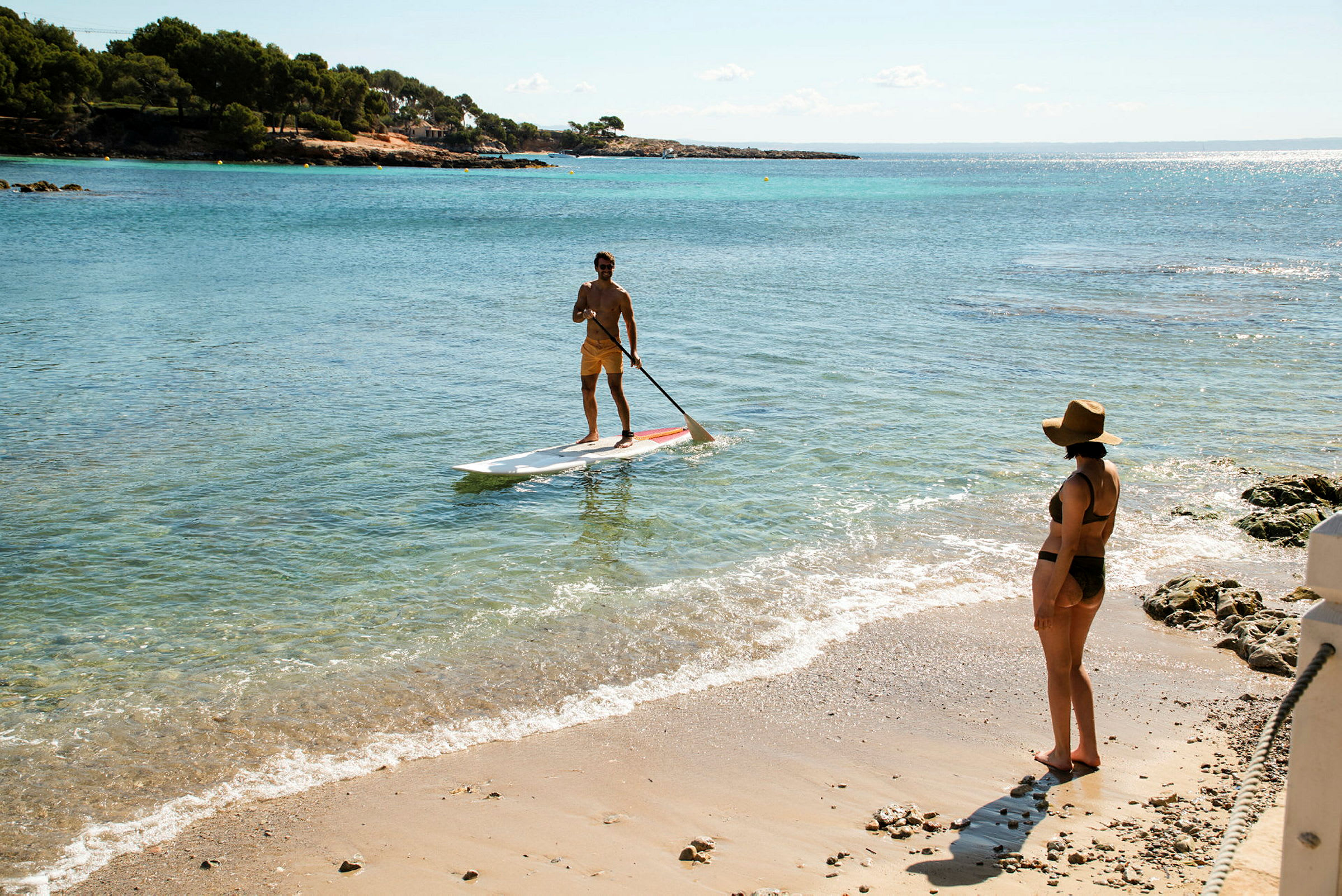 En person står på et paddleboard i havet nær stranden, mens en annen person står på sandstranden og ser mot vannet. (AI-generert tekst)