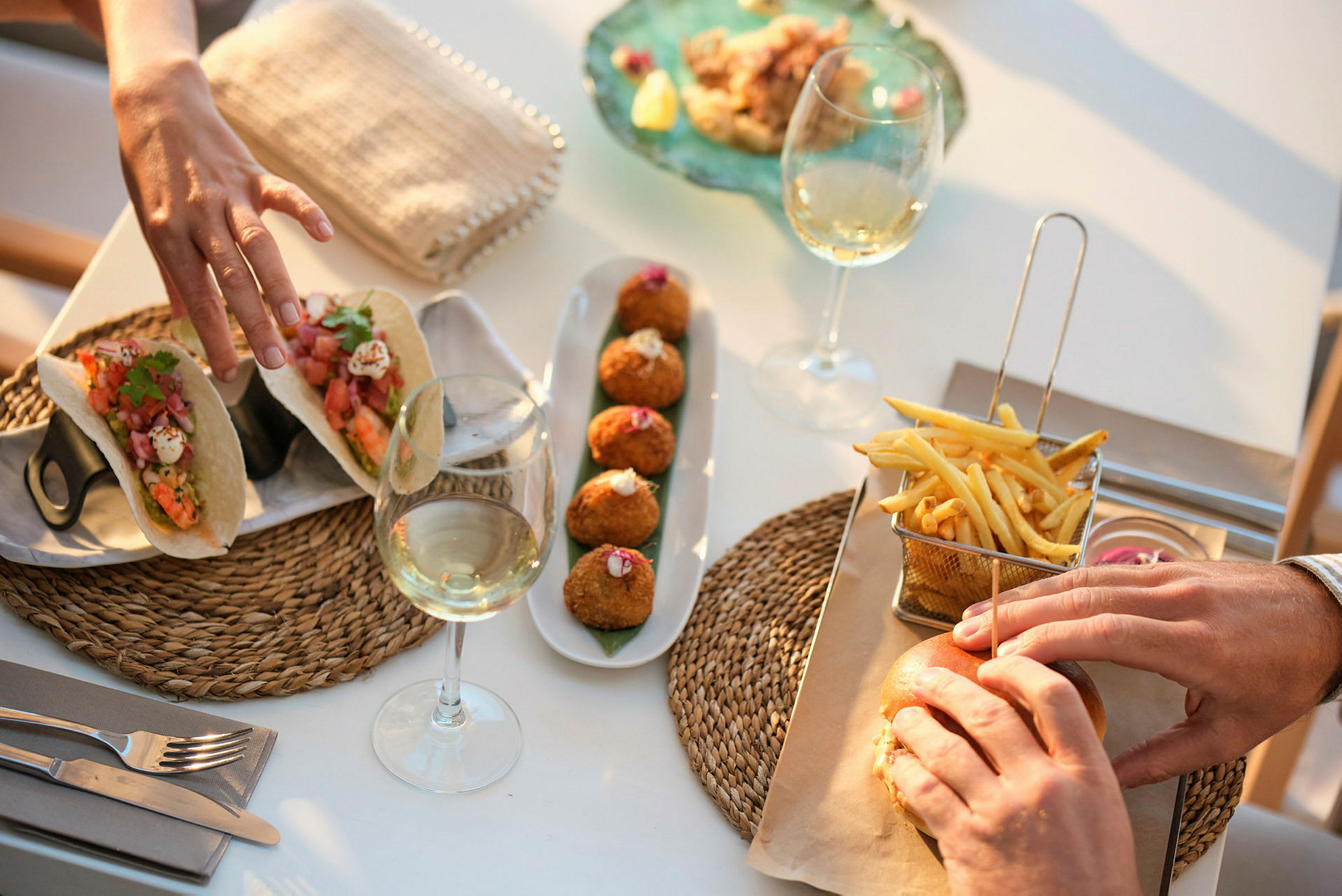 Nærbilde av en bordssituasjon med mat og drikke, inkludert tacos, croquetas, pommes frites, en hamburger og vinglass. (AI-generert tekst)