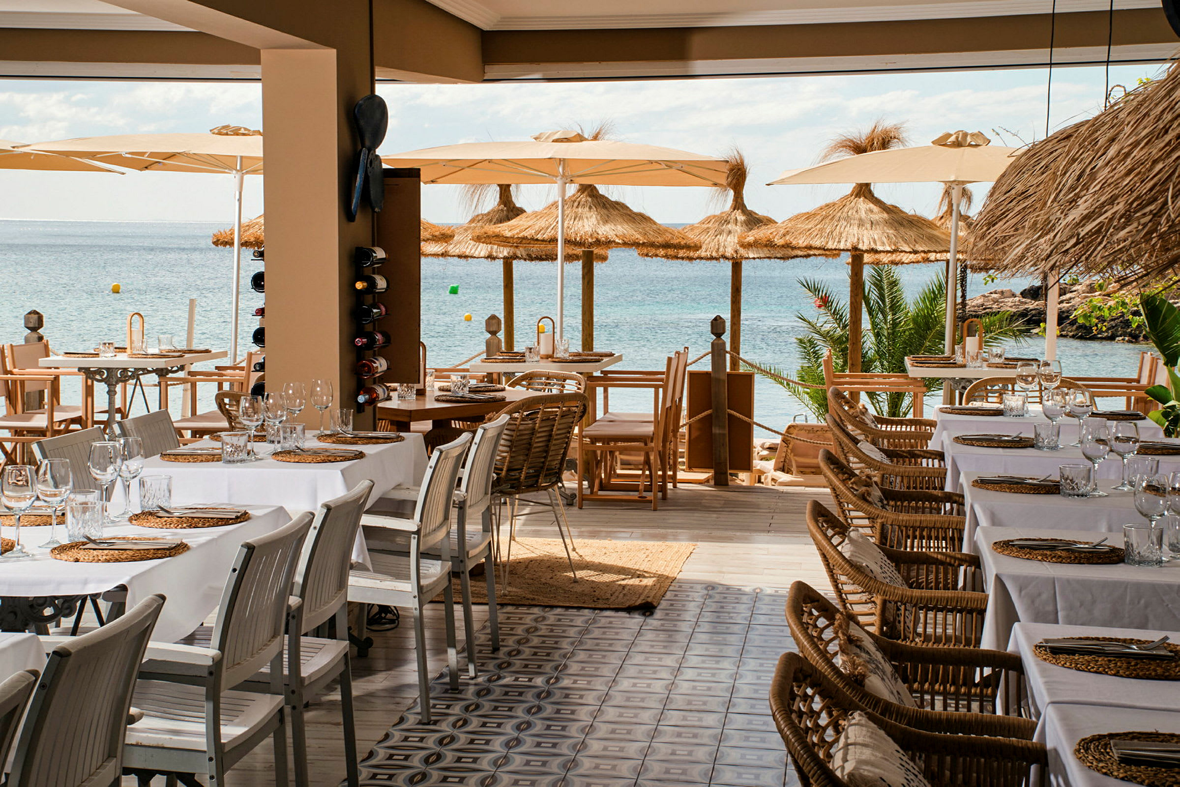 Bilde av en restaurant med dekkede bord og utsikt over havet, parasoller og strandfront. (AI-generert tekst)