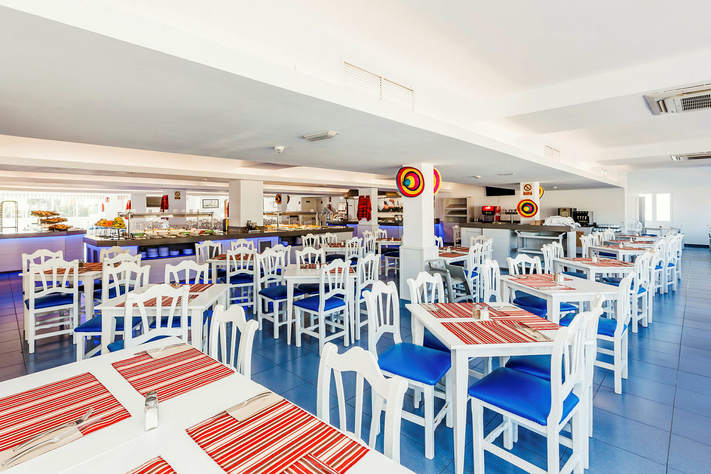 Interiør av en restaurant med hvite bord og stoler med blå puter og røde stripete bordbrikker. (AI-generert tekst)