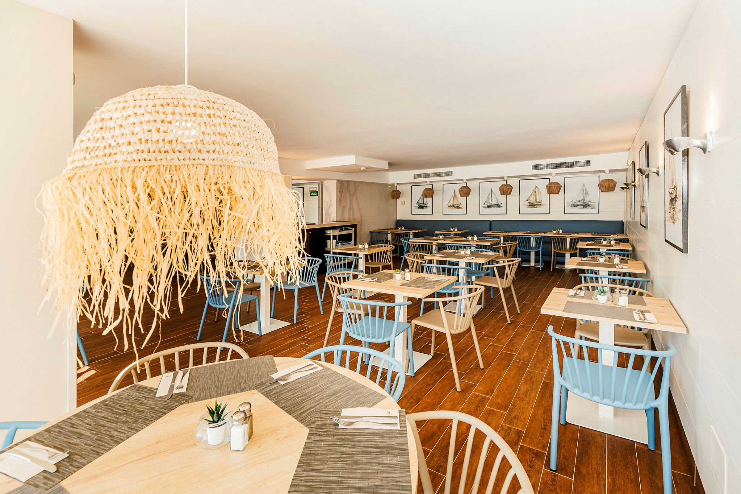Interiør av en restaurant med trebord, blå og beige stoler samt veggdekorasjoner med båtmotiver. (AI-generert tekst)