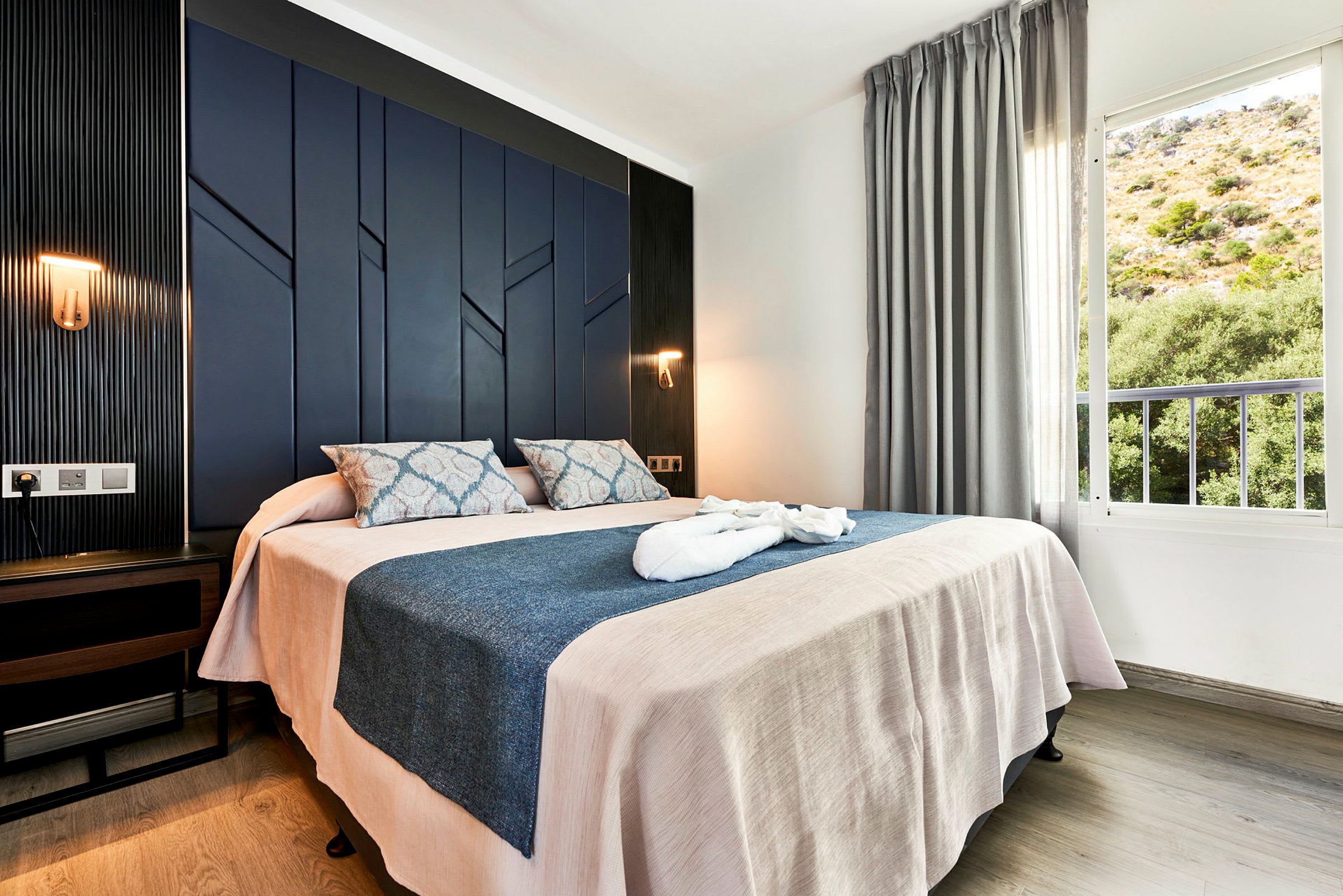 Hotellrom med dobbeltseng, mørkeblå panelvegg og vindu med utsikt over et frodig område. (AI-generert tekst)