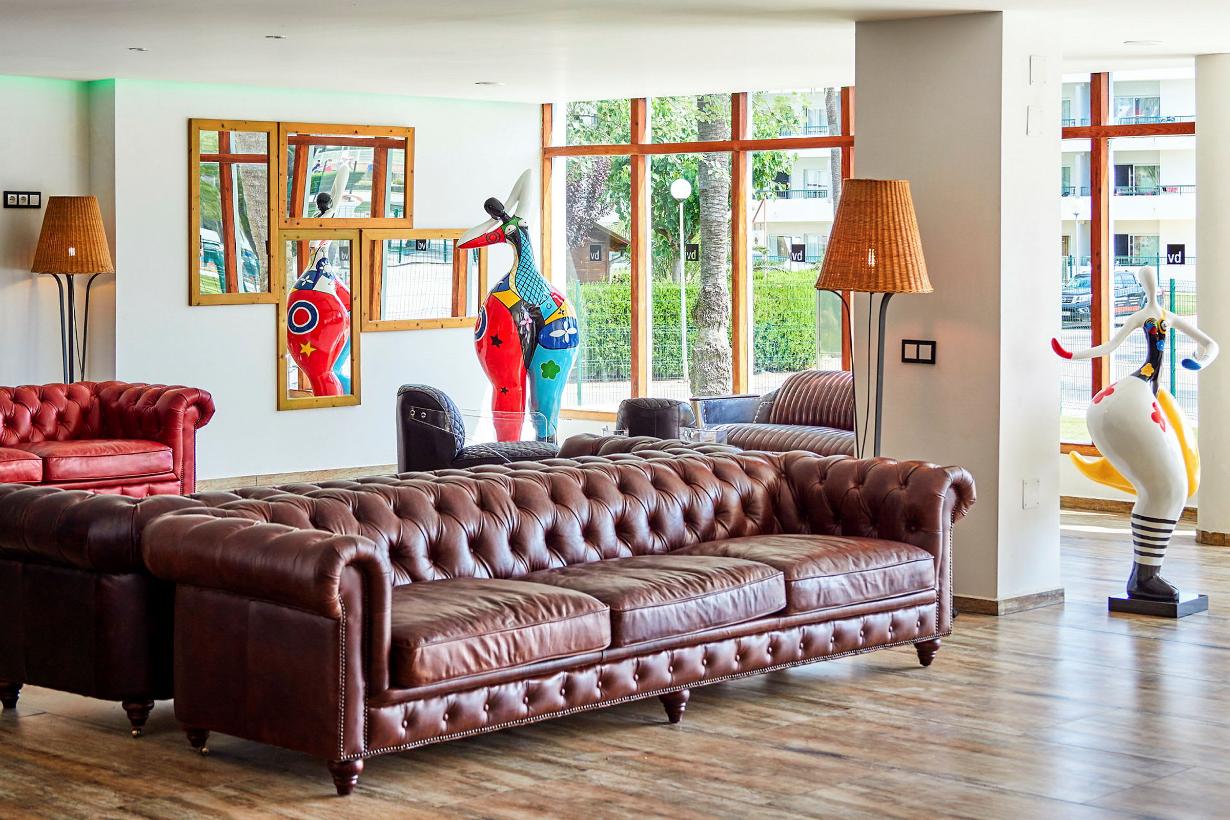 En hotellobby med en brun chesterfield-sofa, dekorative skulpturer og store vinduer med utsikt. (AI-generert tekst)