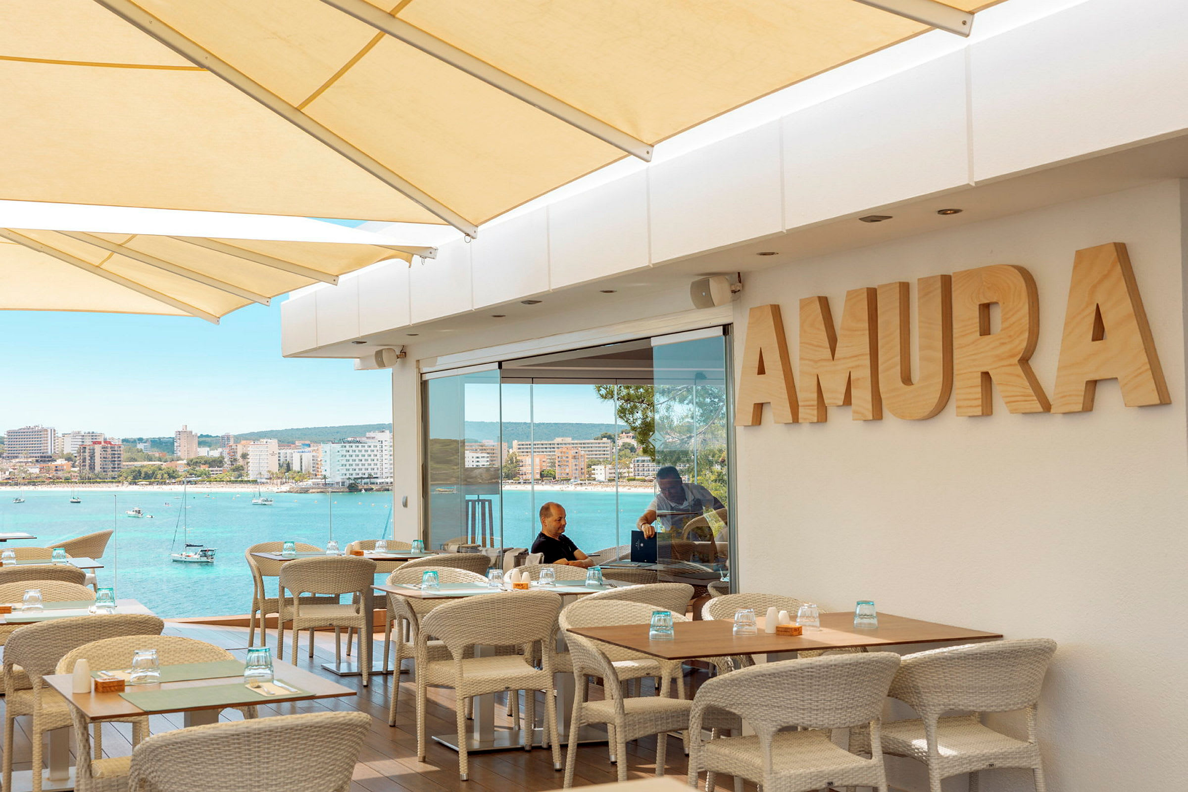 Bilde av en uterestaurant med bord og stoler, et skilt med teksten 'AMURA', og utsikt over blått vann og en byhorisont i bakgrunnen. (AI-generert tekst)