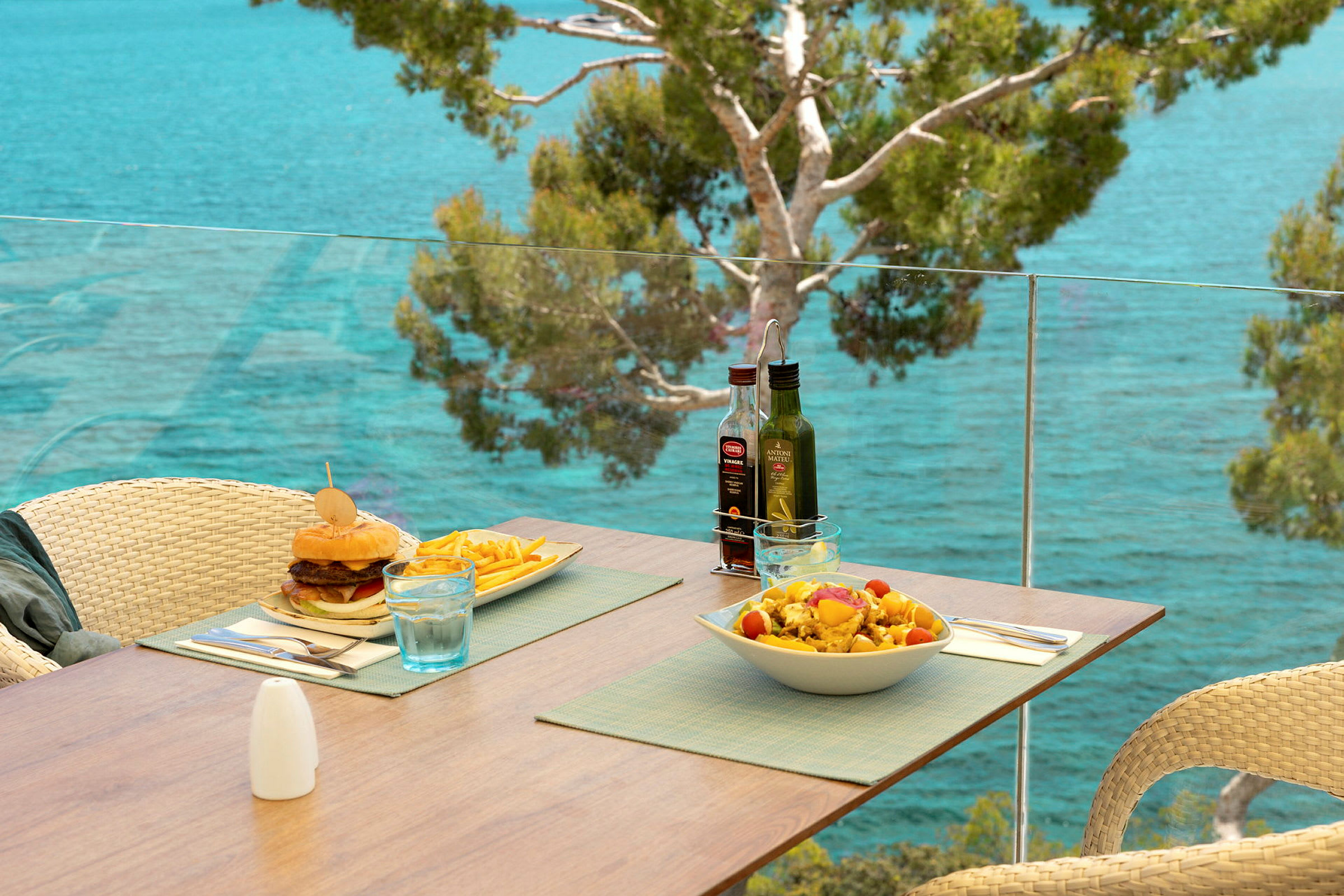 En terrasse med utsikt over havet, hvor et bord er dekket med en hamburger, pommes frites og en salat. (AI-generert tekst)