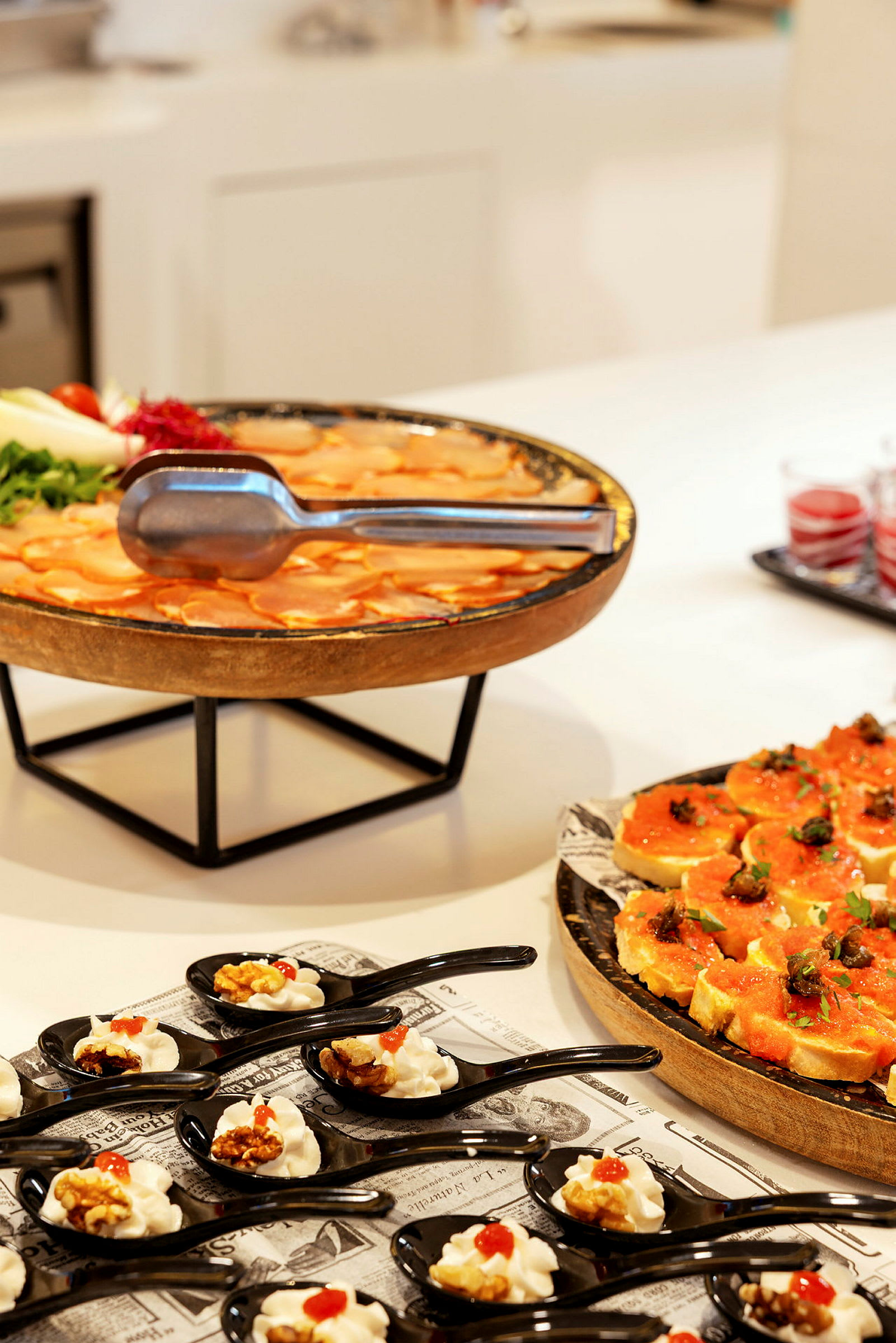 En buffet med ulike retter, inkludert små porsjoner servert i svarte skjeer, en tallerken med oransje farget mat og dekorerte brødskiver. (AI-generert tekst)