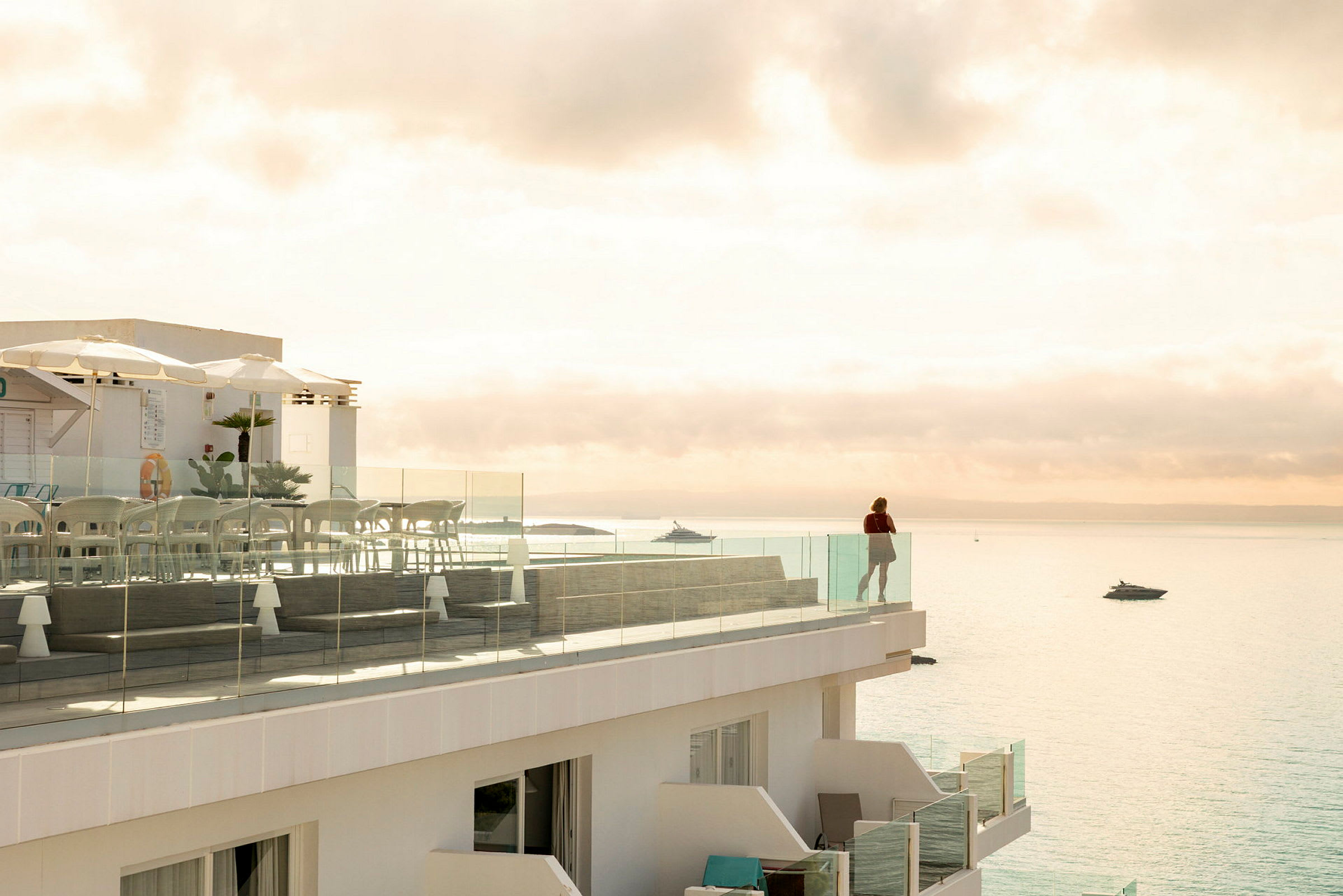 En terrasse med glassrekkverk med utsikt over havet. På terrassen står det stoler og bord, og en person står ved rekkverket og ser utover vannet. (AI-generert tekst)