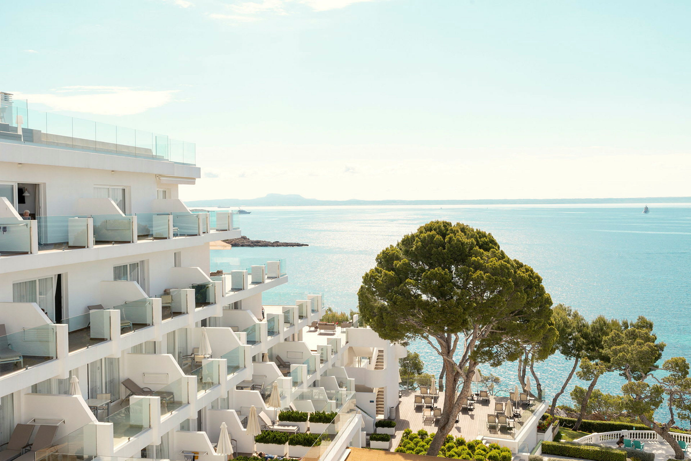 Hotellbygning ved kysten med utsikt over havet og en terrasse med bord og stoler samt trær. (AI-generert tekst)