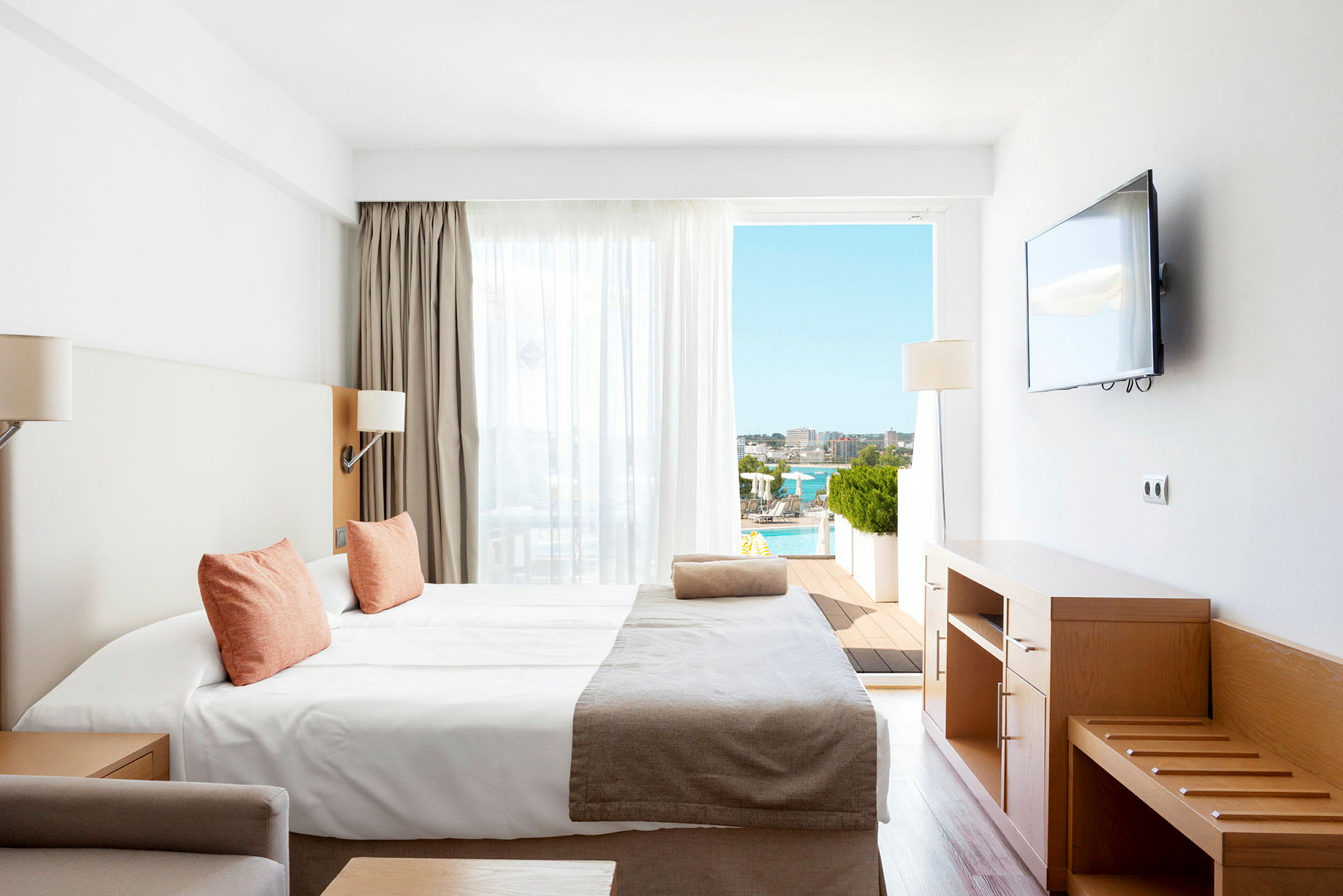Et hotellrom med en seng, tremøbler, en veggmontert TV og utsikt over en solrik terrasse med basseng. (AI-generert tekst)