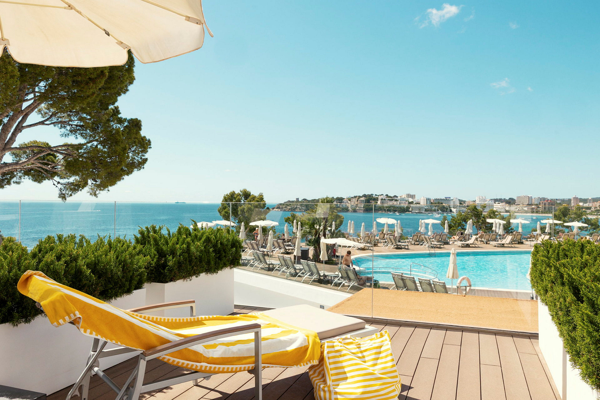 En solseng med gulgul håndkle og strandveske står på en terrasse med utsikt over et basseng og havet. (AI-generert tekst)