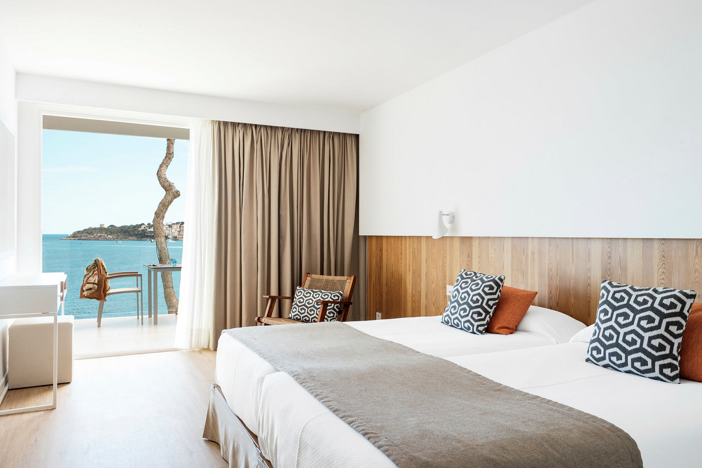 Et hotellrom med moderne detaljer, en dobbeltseng, rustikke puter, en sitteplass og en balkong med utsikt over havet. (AI-generert tekst)