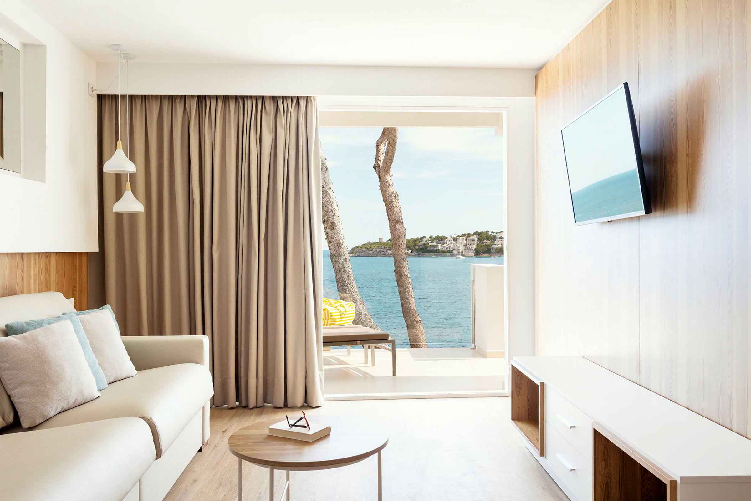 En lys stue med en sofa, bord og TV. Vinduet viser utsikt over havet og noen trær. (AI-generert tekst)