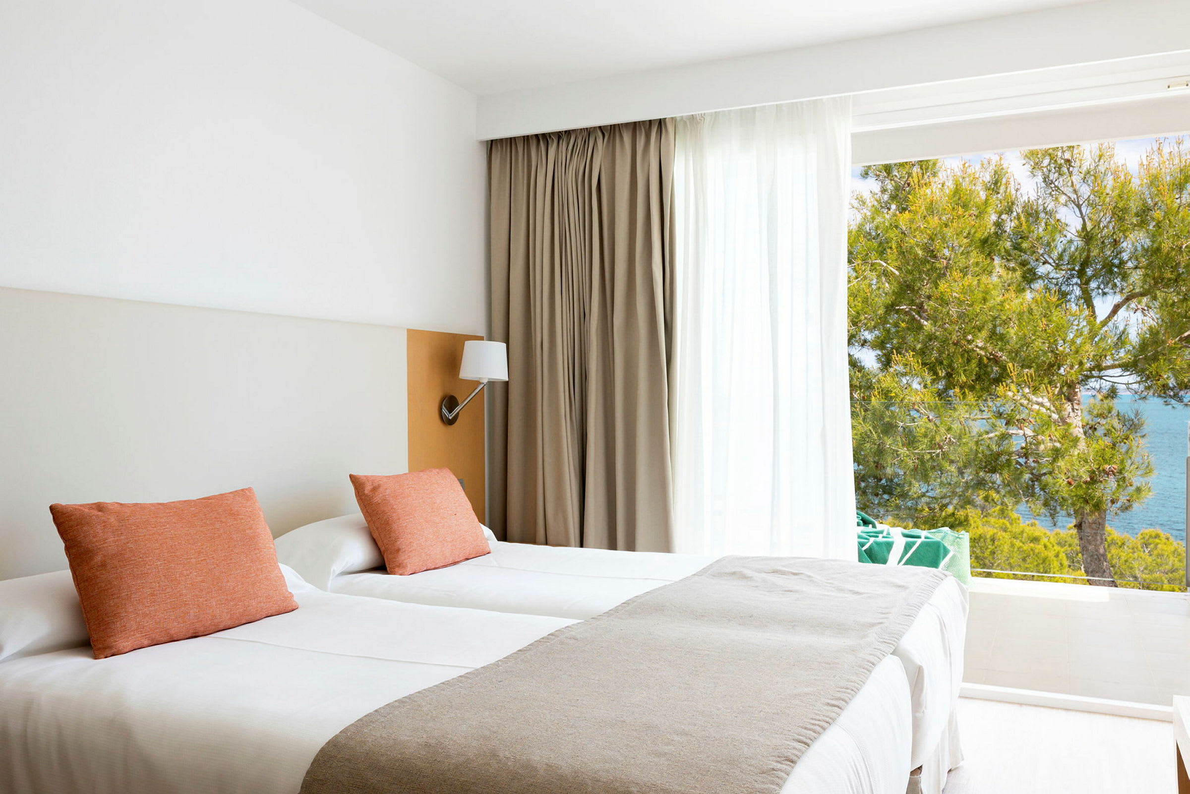 Et lyst og moderne hotellrom med to enkeltsenger, pynteputer og utsikt mot havet gjennom store vinduer. (AI-generert tekst)